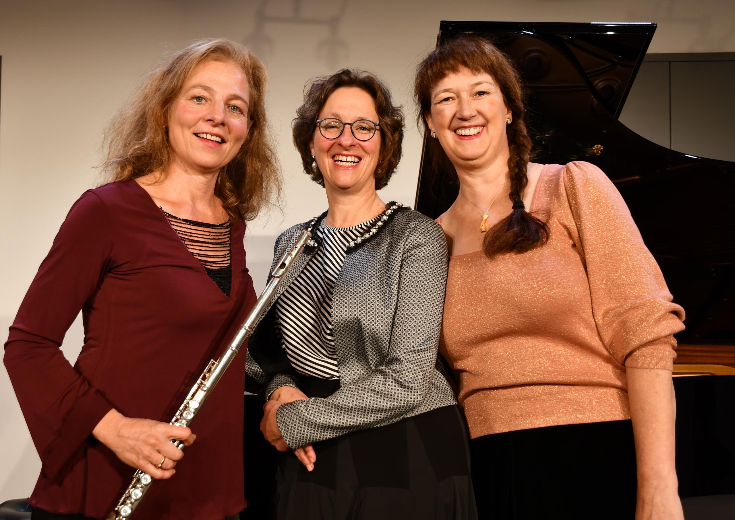 Three ladies lean against the grand piano and laugh into the camera. The lady on the left has a flute in her hand.