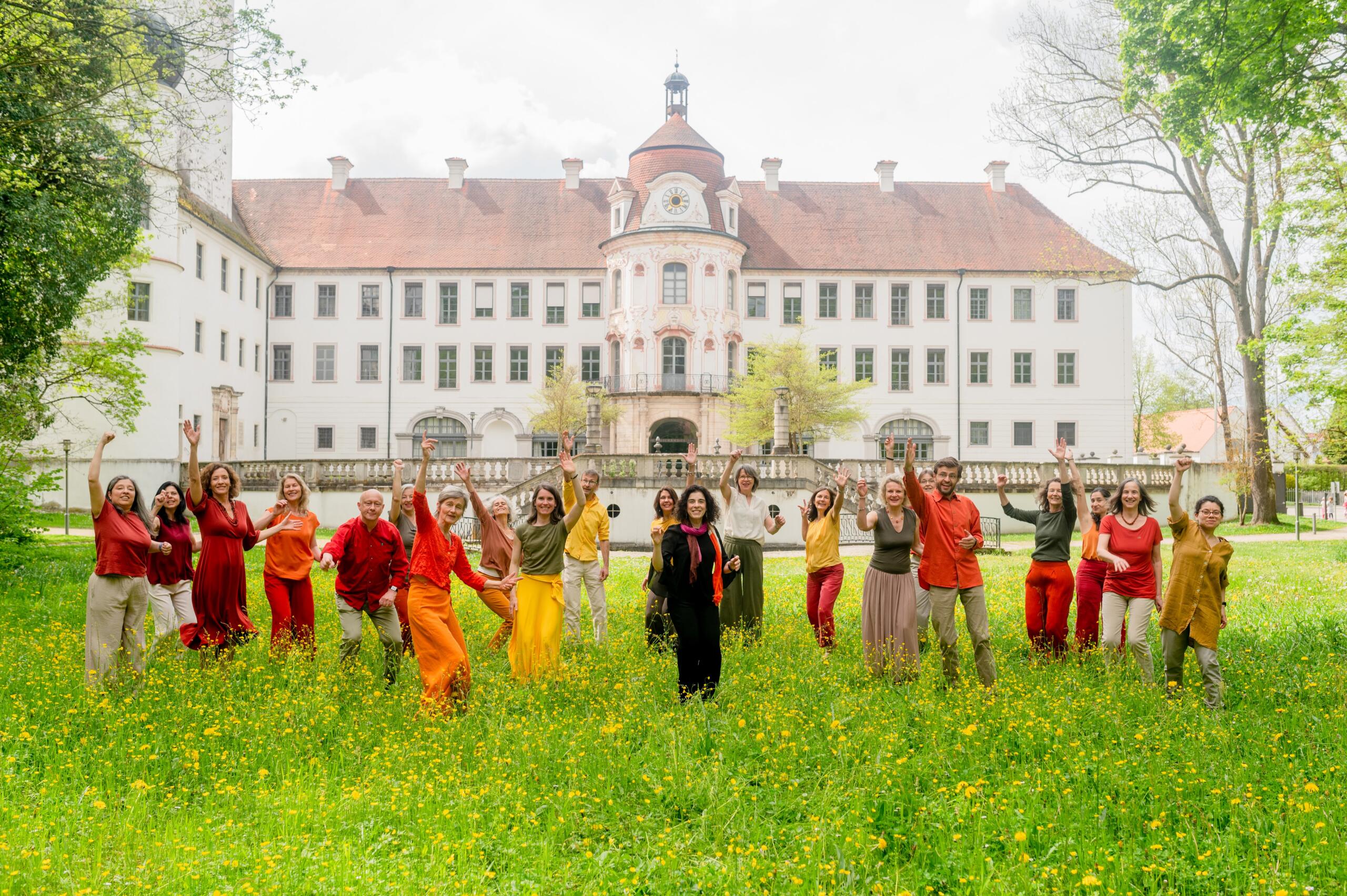 People in colourful clothes on a green meadow, waving. A Schliss can be seen in the background.
