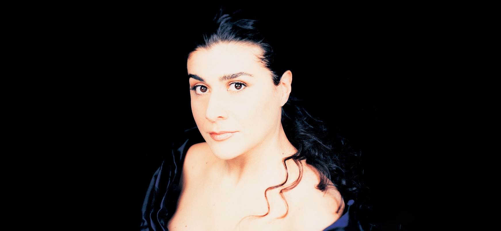 Portrait shot of singer Cecilia Bartoli against a black background. She looks directly into the camera.