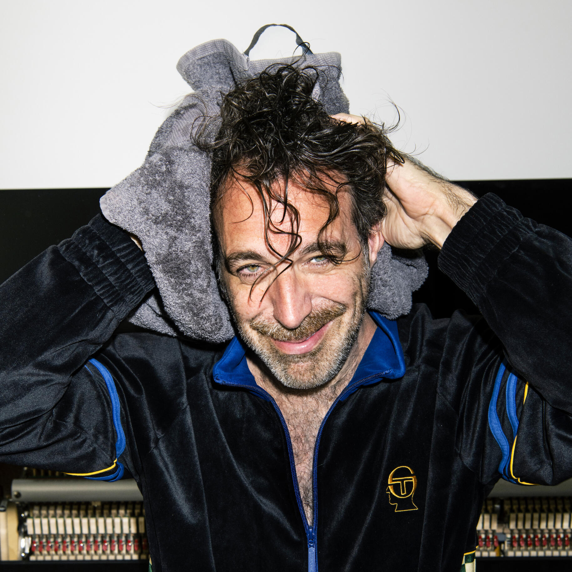Ein Porträt des Musikers und Kabarettisten Chilly Gonzales. Er trägt einen Trainingsanzug aus Samt, sitzt vor einem Klavier und rubbelt sich mit einem Handtuch die lockigen Haare trocken.