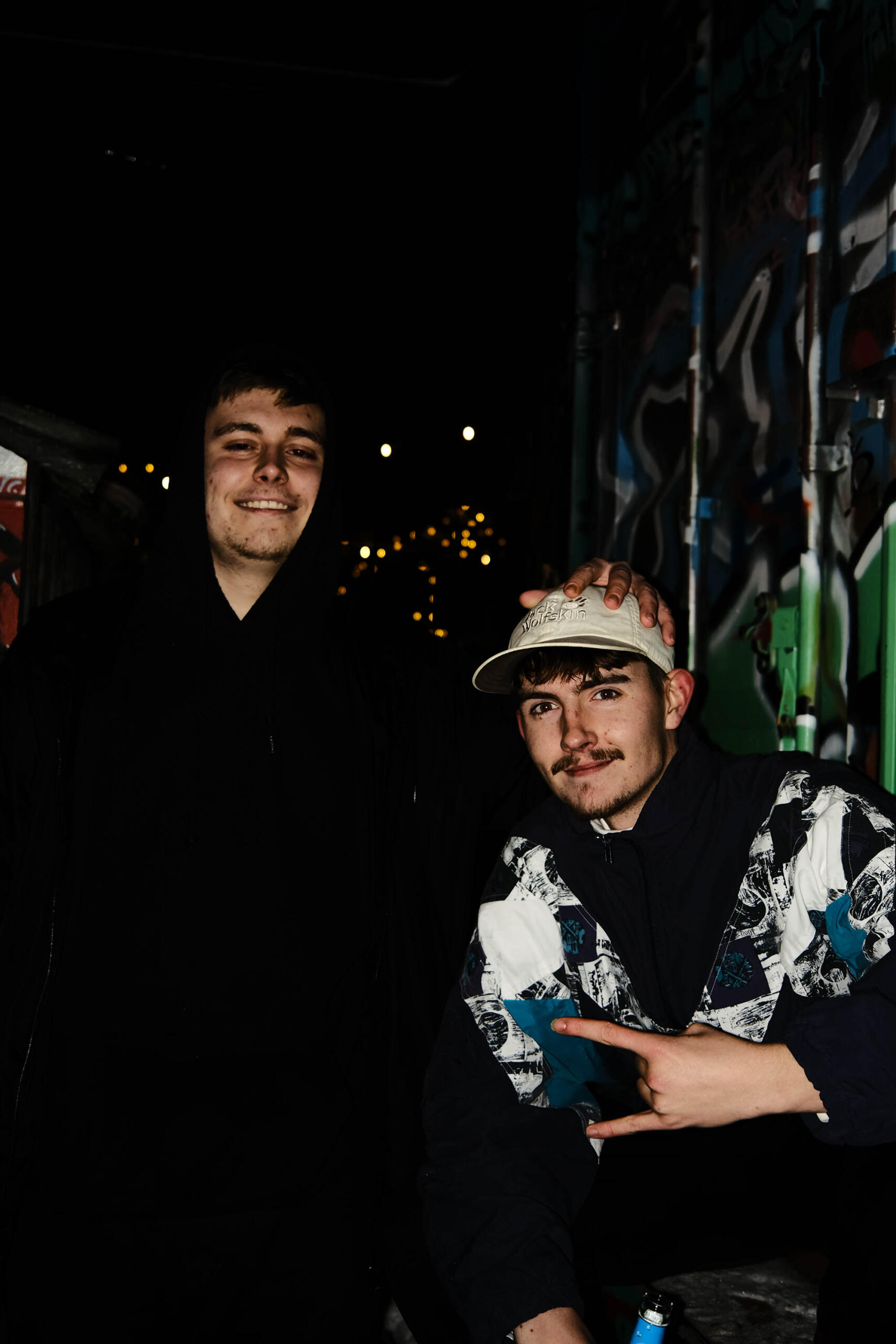 Two young men next to a graffiti wall at night.