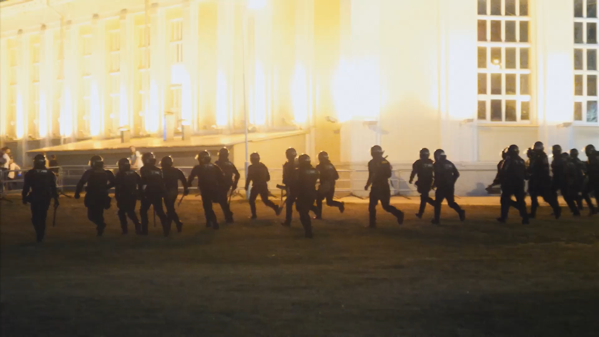 A crowd of masked policemen run towards a lit-up building.