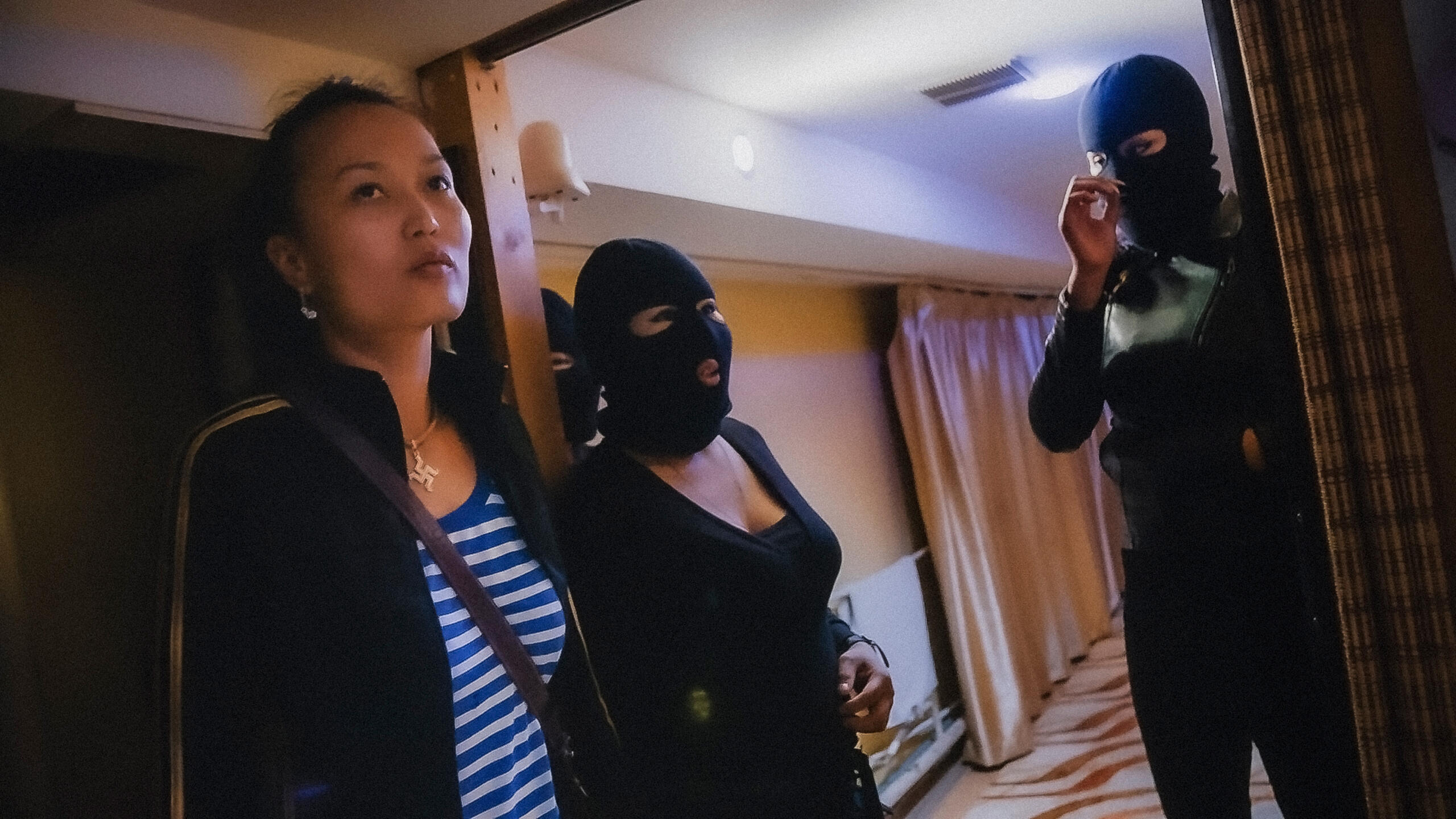Eine Frau in einem Streifenshirt und einer Trainingsjacke steht mit zwei vermummten Frauen in einem Hotelzimmer.