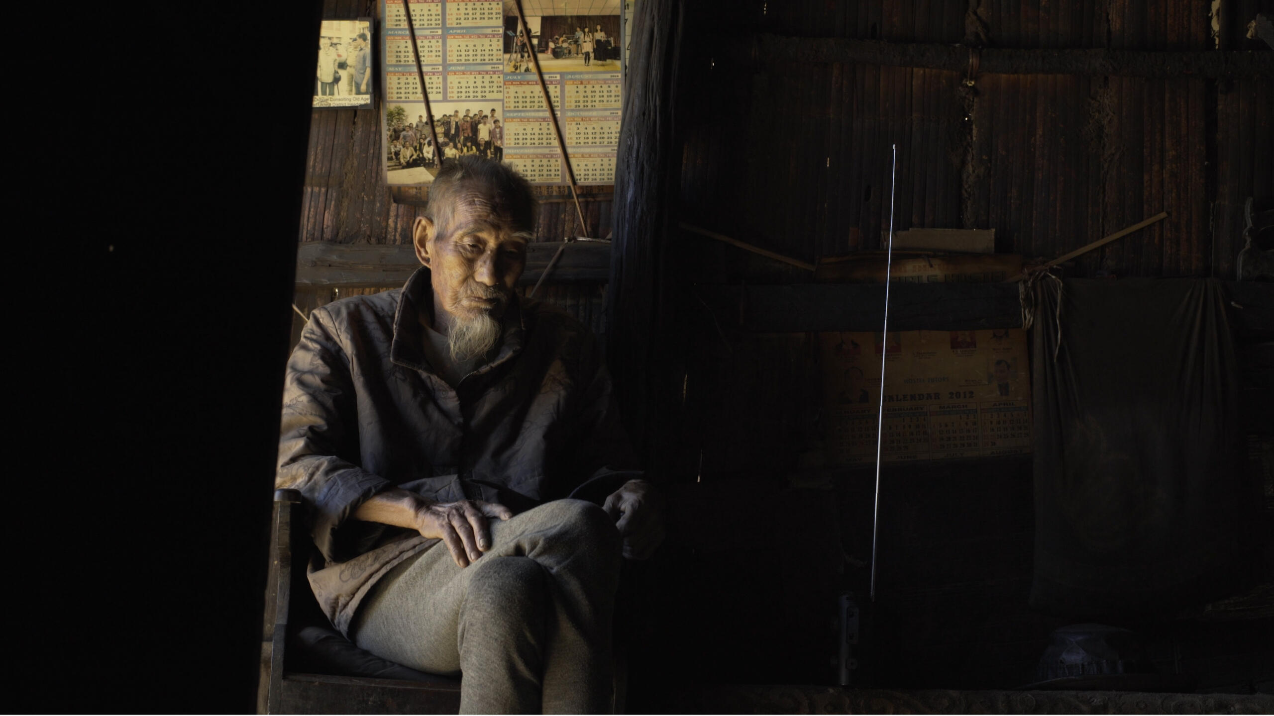 An old man sits in a dark room, wrapped in a blanket and covered in dirt.