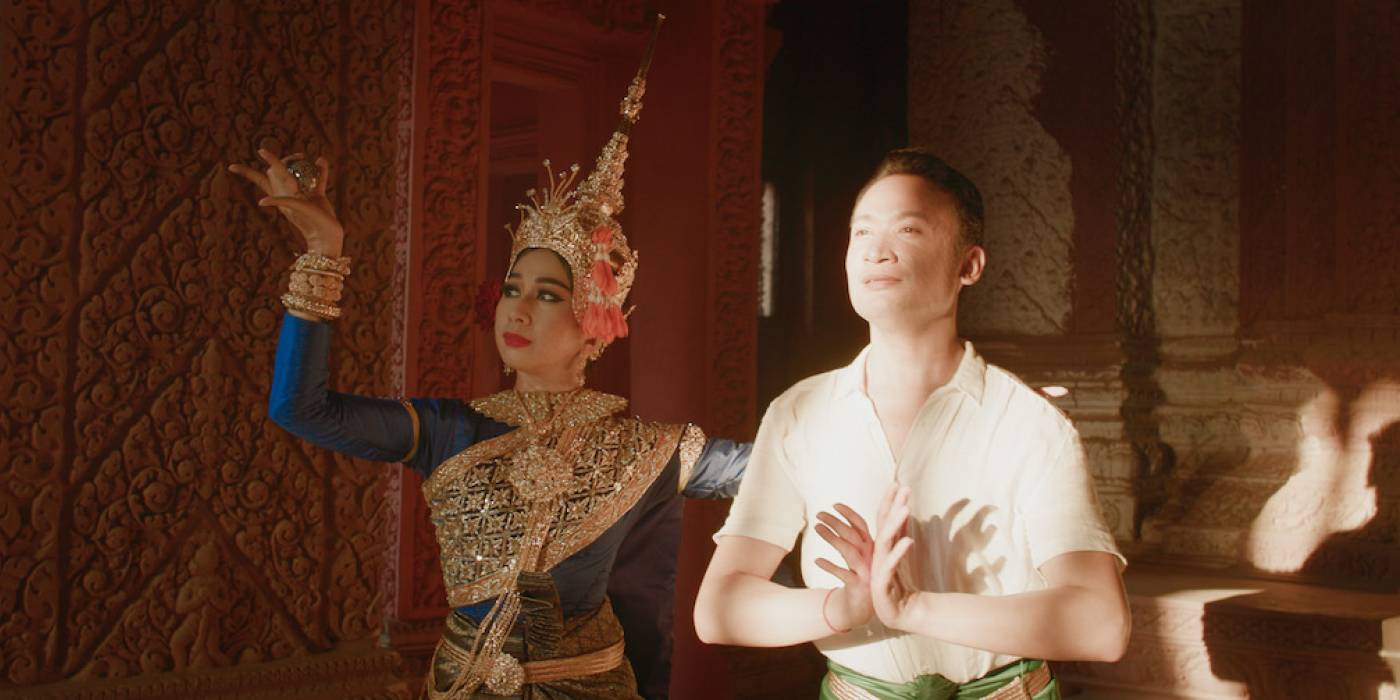 A man in a simple outfit and a woman wearing a golden dress and lots of golden jewellery dance in a room.