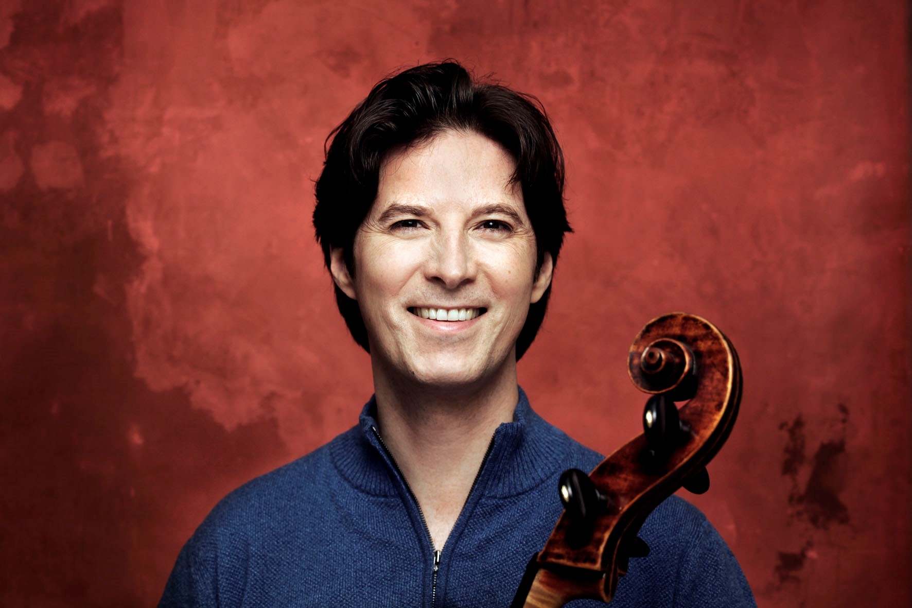 Portraitaufnahme des Cellist Daniel Müller-Schott. Er steht vor einer roten Wand und trägt einen blauen Pullover.