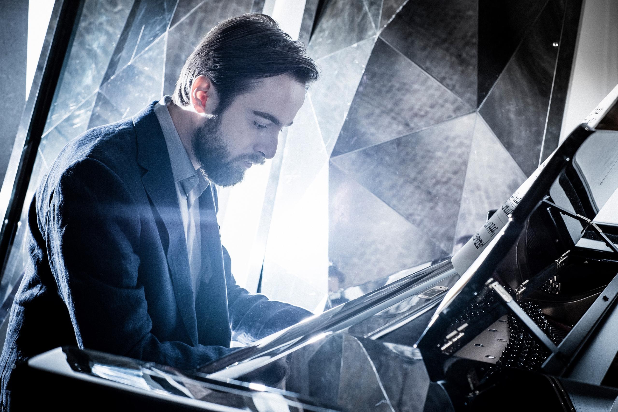The pianist Daniil Trifonov sits at a black grand piano in front of a background consisting of many small, metallic reflecting surfaces.