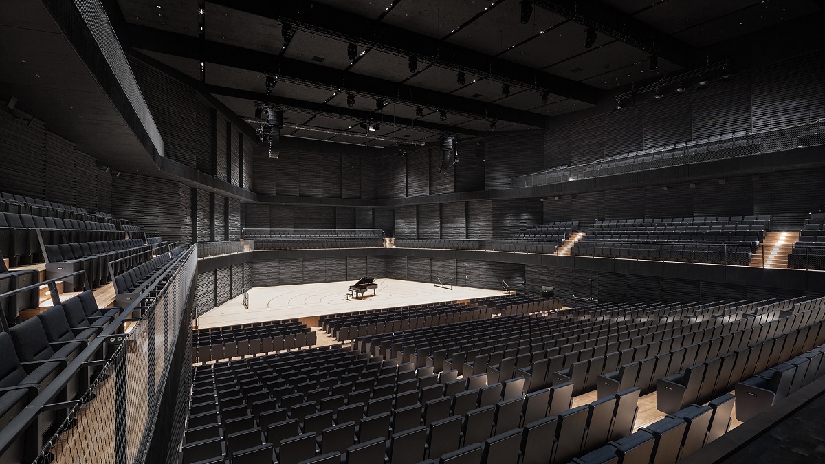 Empty concert hall with black wood paneling and light stage on which stands a grand piano