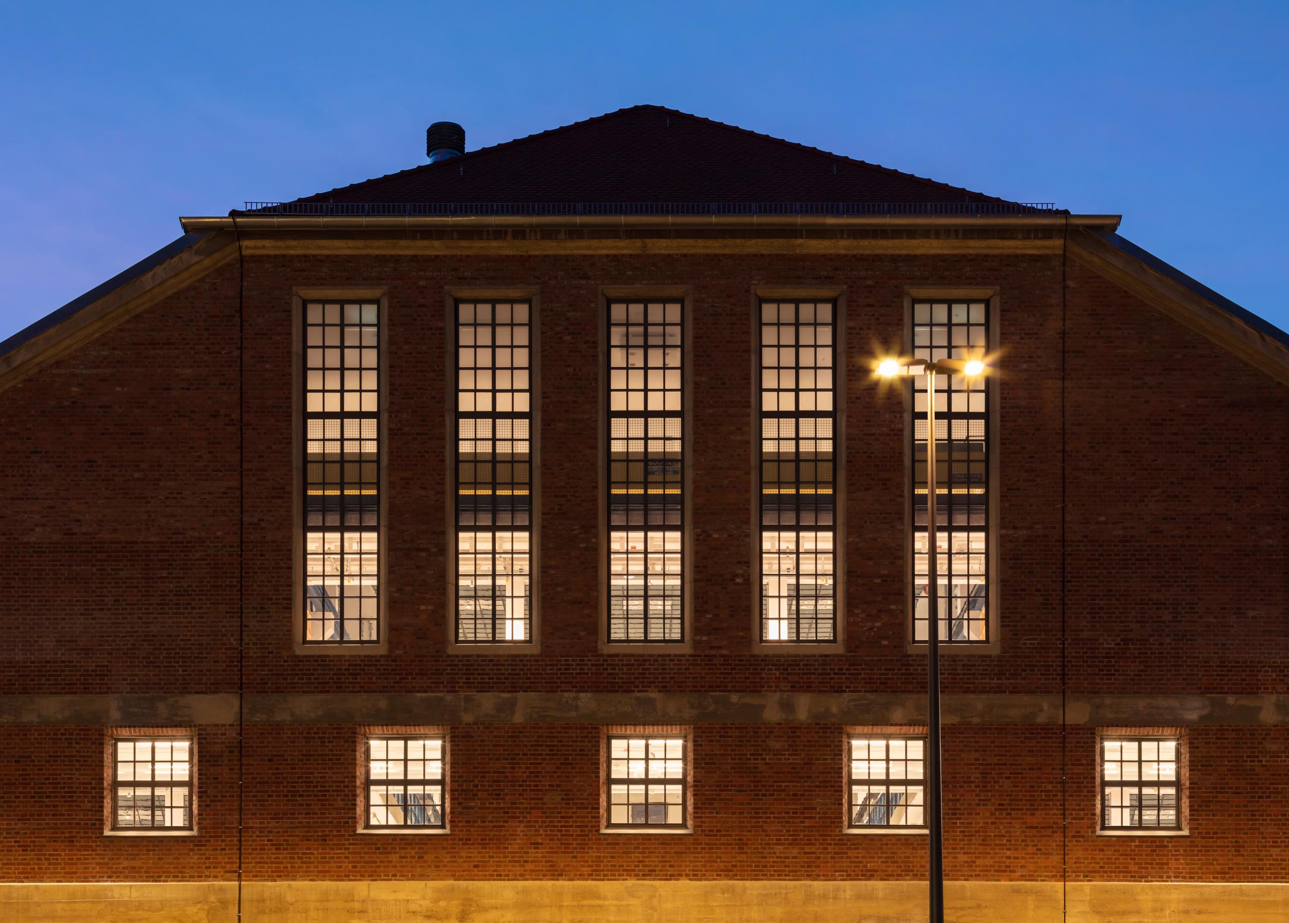 Stirnseite der Backstein-Halle E abends beleuchtet, mit 5 hohen Fenstern und darunter 5 kleinen quadratischen, Straßenlatererne