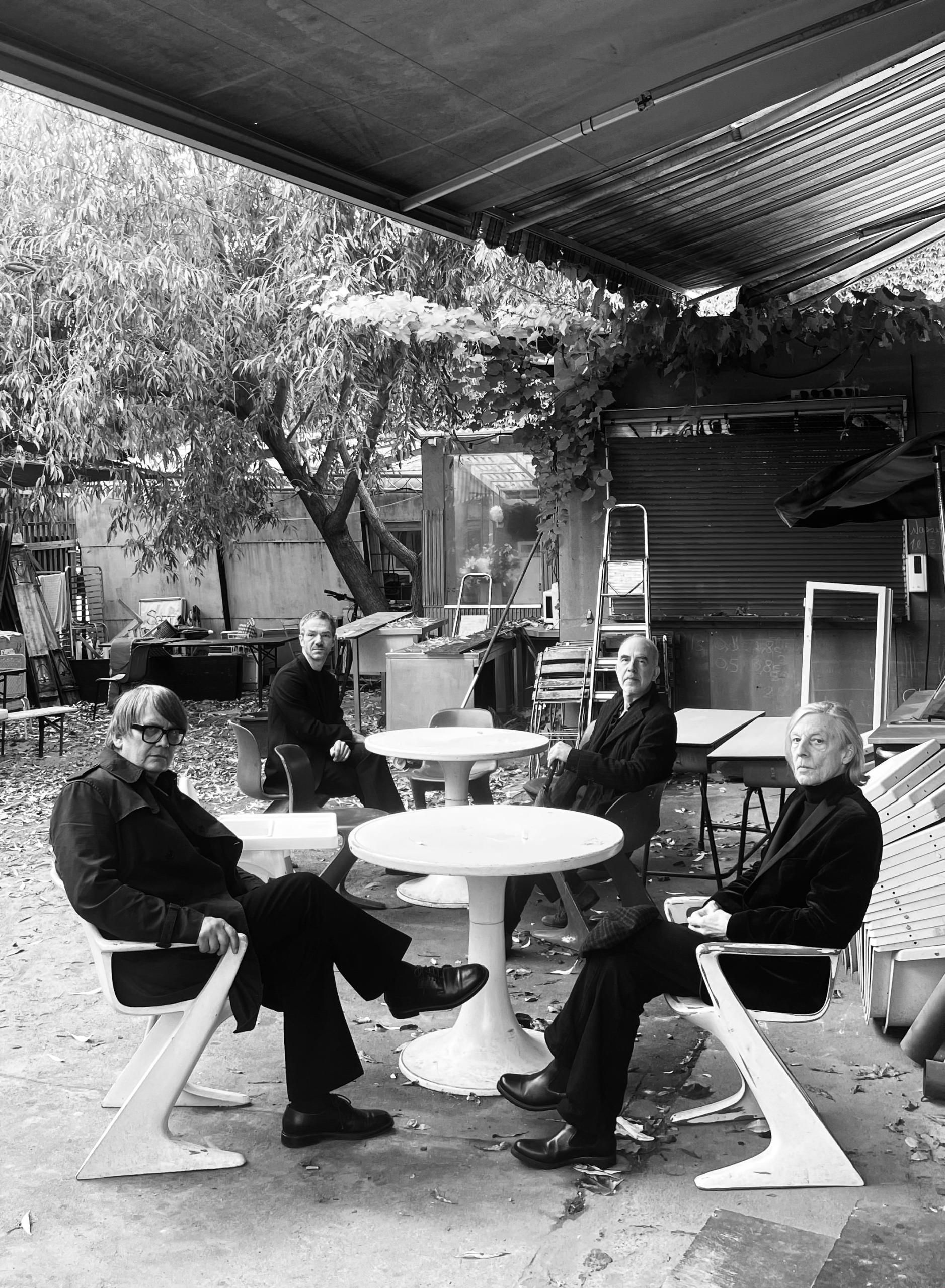 The 4 musicians of the band Element of Crime are sitting on chairs around two small tables under an eaves. A garden can be seen in the background.