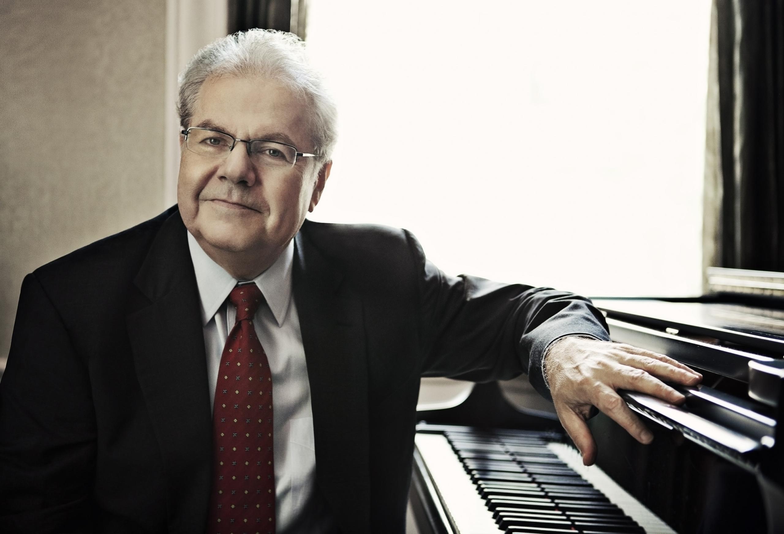 Pianist Emanuel Ax sits next to a black grand piano and looks into the camera