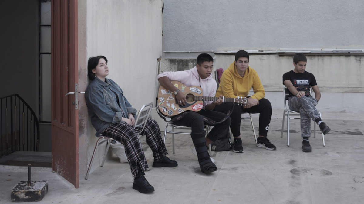 Vier Jugendliche sitzen auf einer Terrasse. Einer hält eine Gitarre in der Hand. Links neben ihnen ist eine offene Tür zu sehen, die in ein Treppenhaus führt.