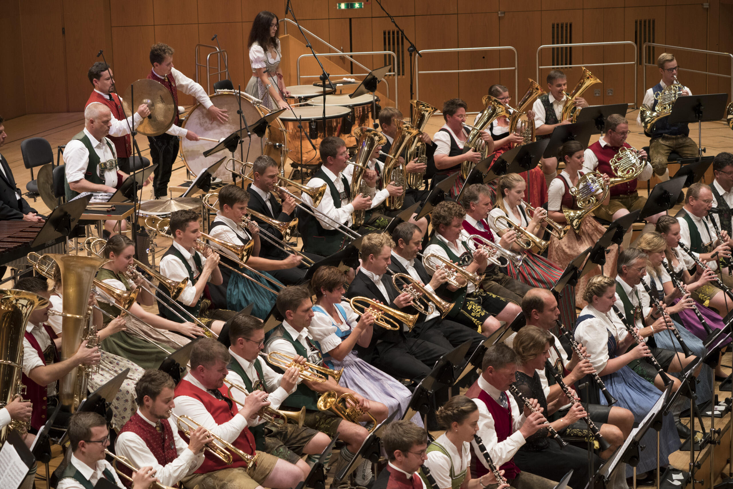 Brass musicians sit in several rows on a stage and play their instruments. They wear Bavarian traditional costumes.