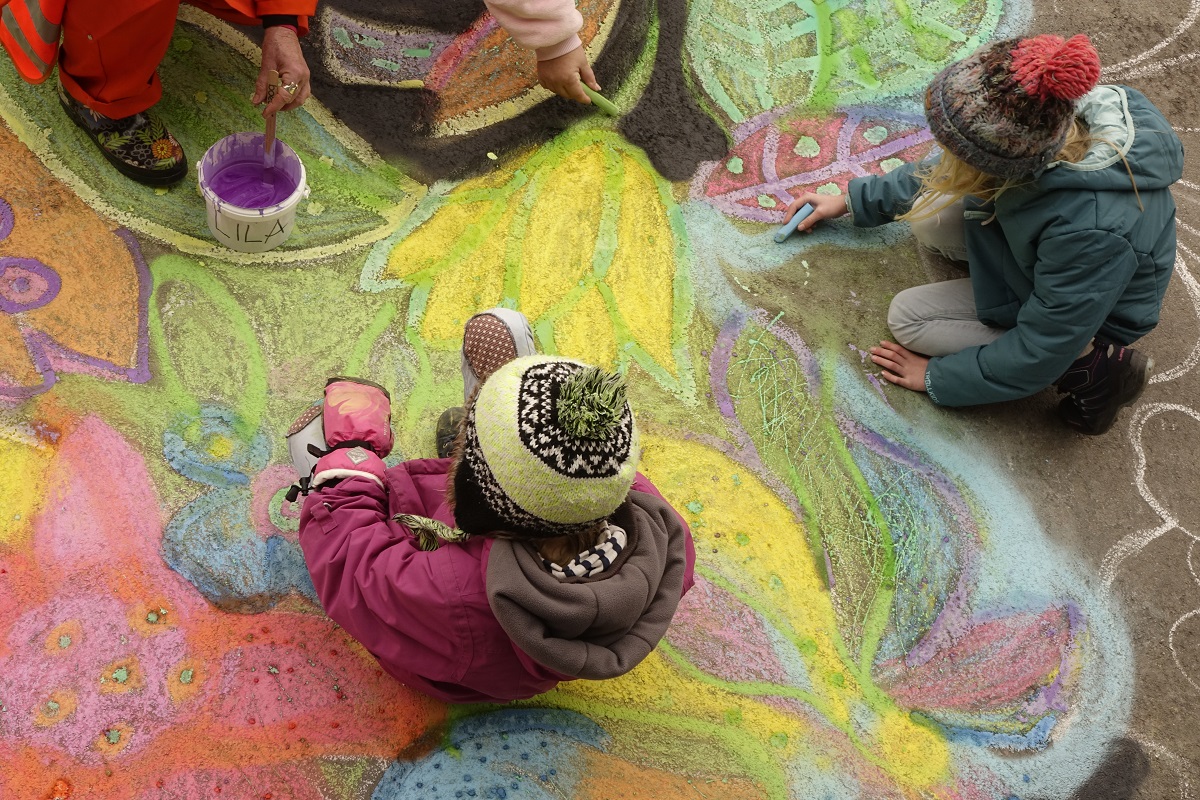 Kinder mit bunten Mützen von oben fotografiert. Sie sitzen auf dem Boden und malen ein Bild aus Kreide.