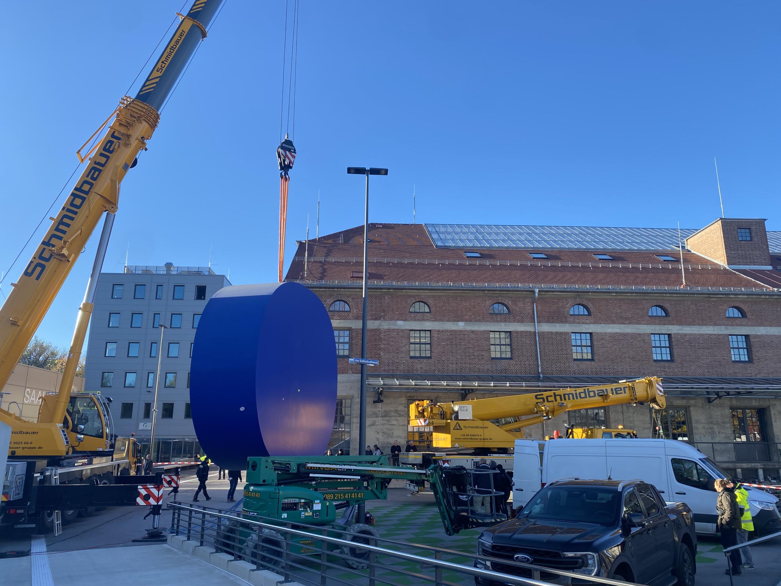 Outside, during the day, a power station in the background, a large, blue, round work of art hanging from a crane in the foreground, a brick building on the left.