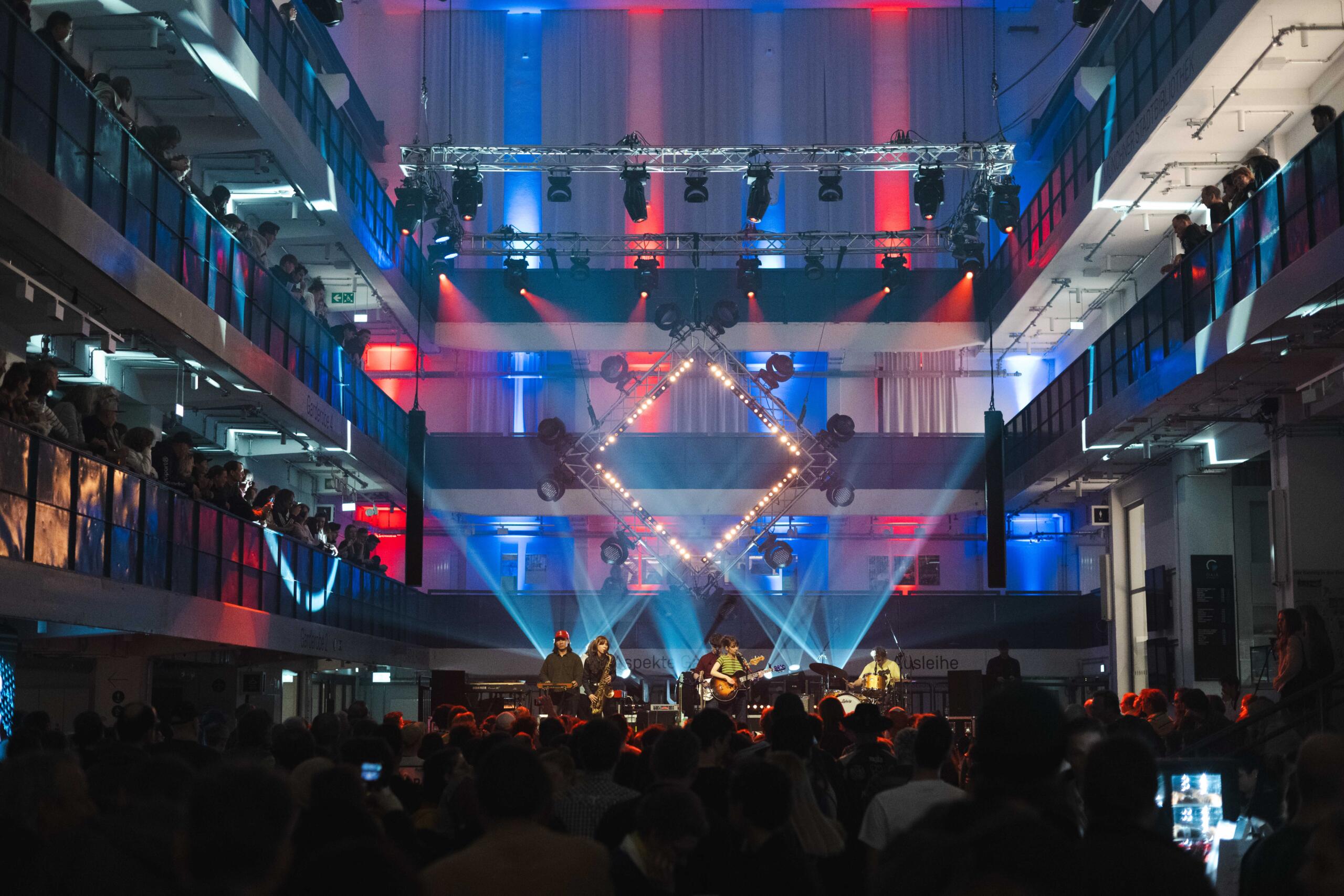 Eine Band spielt vor großem Publikum in der Halle E, die farbig beleuchtet ist und zusätzlich von unten angestrahlt wird 