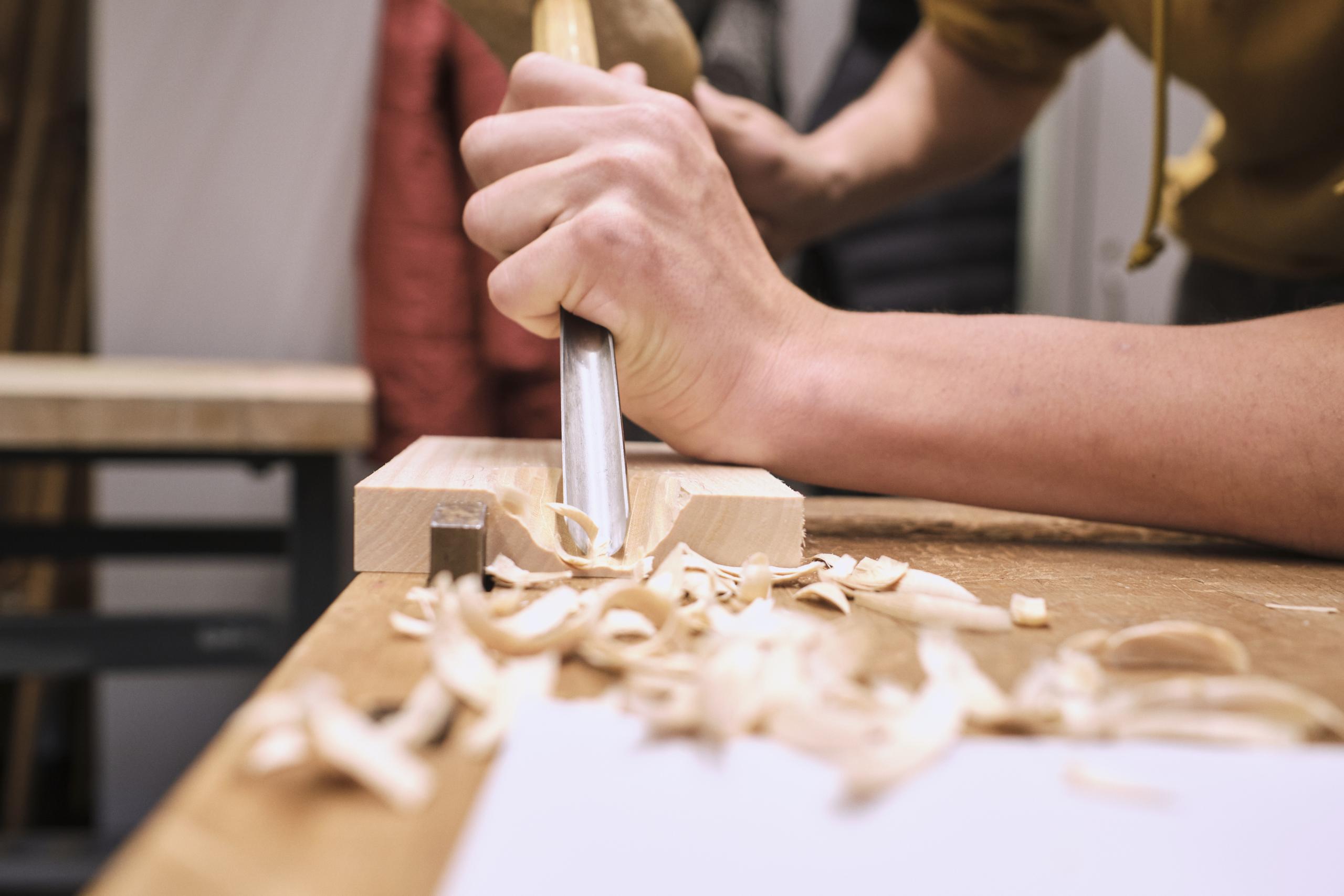 Zwei Hände bearbeiten ein Stück Holz mit Hammer und Meisel