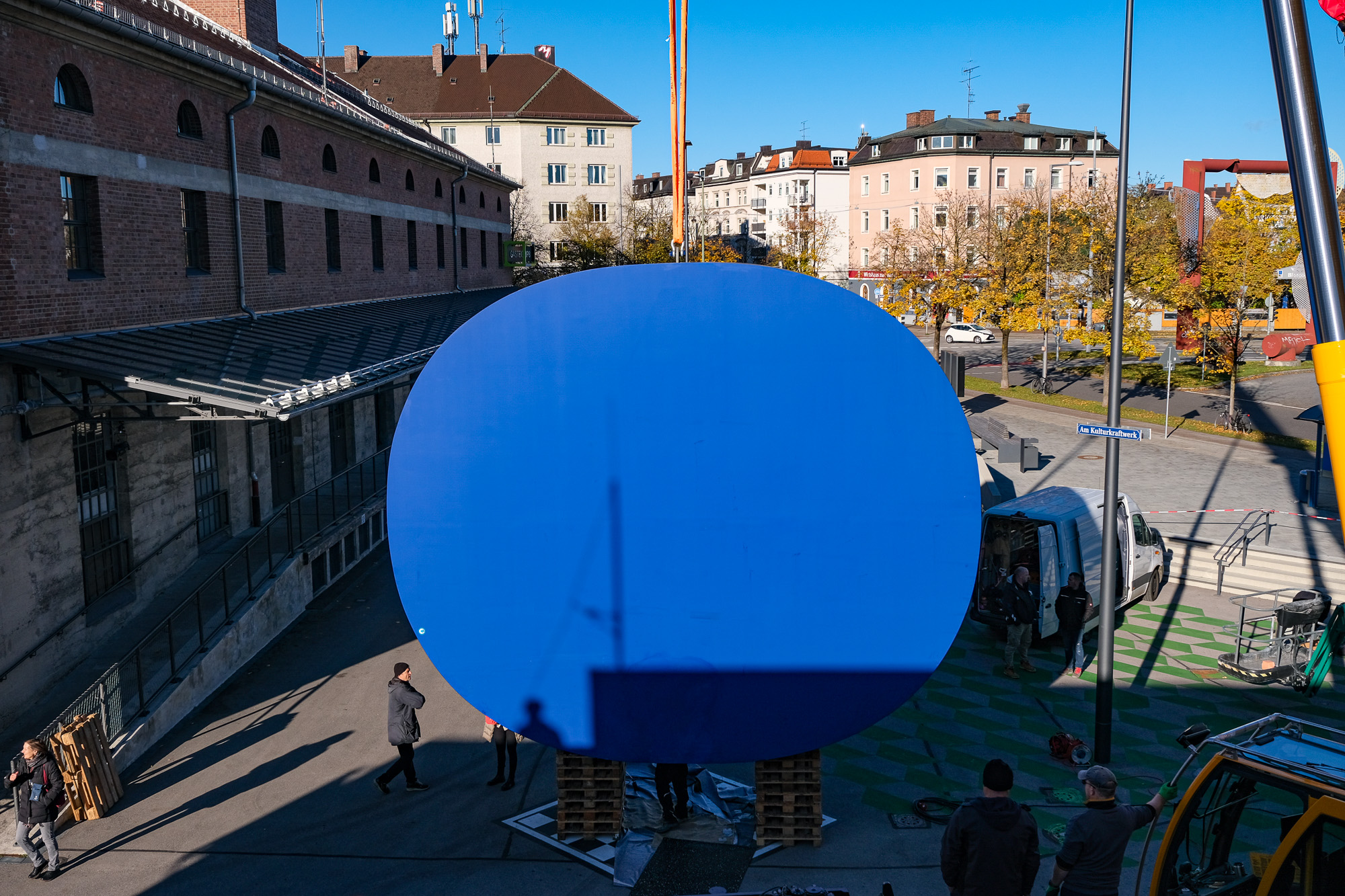 Outdoor area, daytime, view from the roof of Hall X onto "Platz am Kulturkraftwerk", the \"Rounded Blue\" hangs from the crane in front of Hall E.