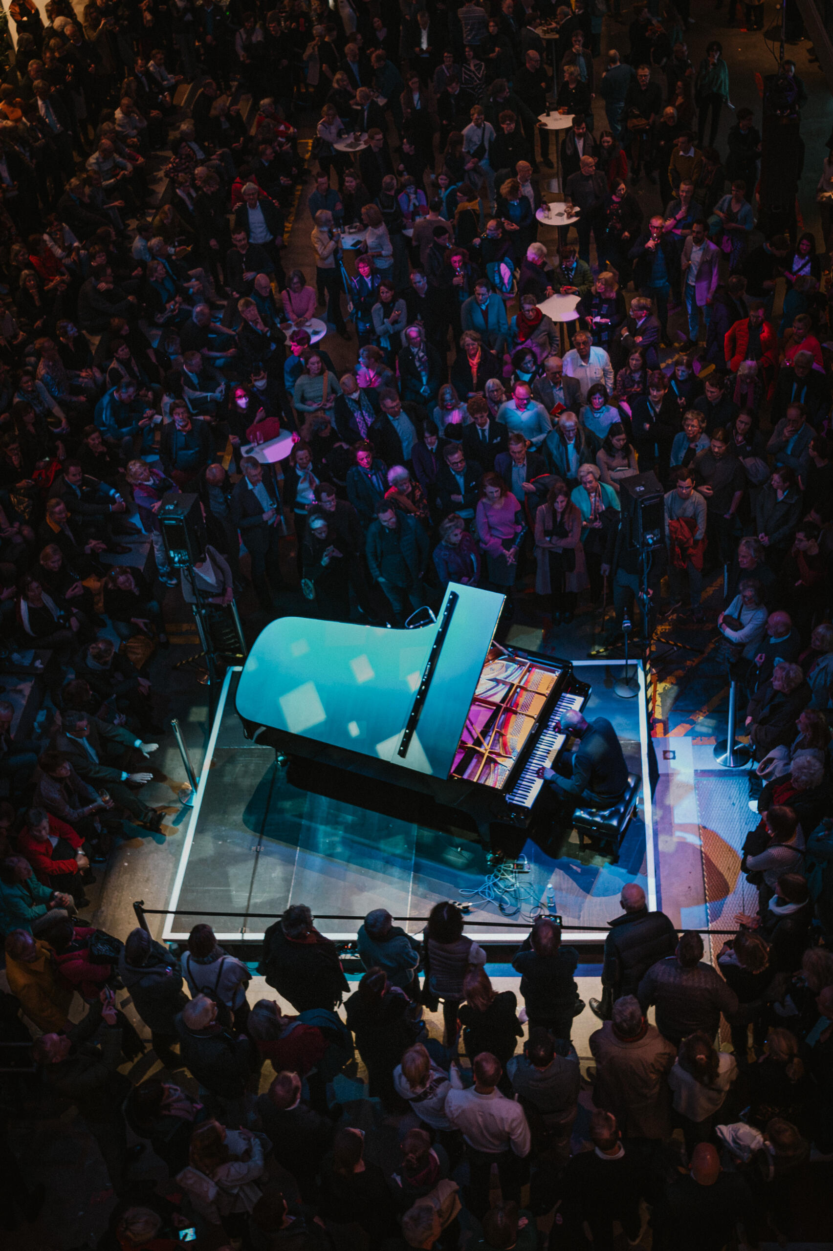 View from above of many people in a hall. They are standing around a grand piano with a pianist.