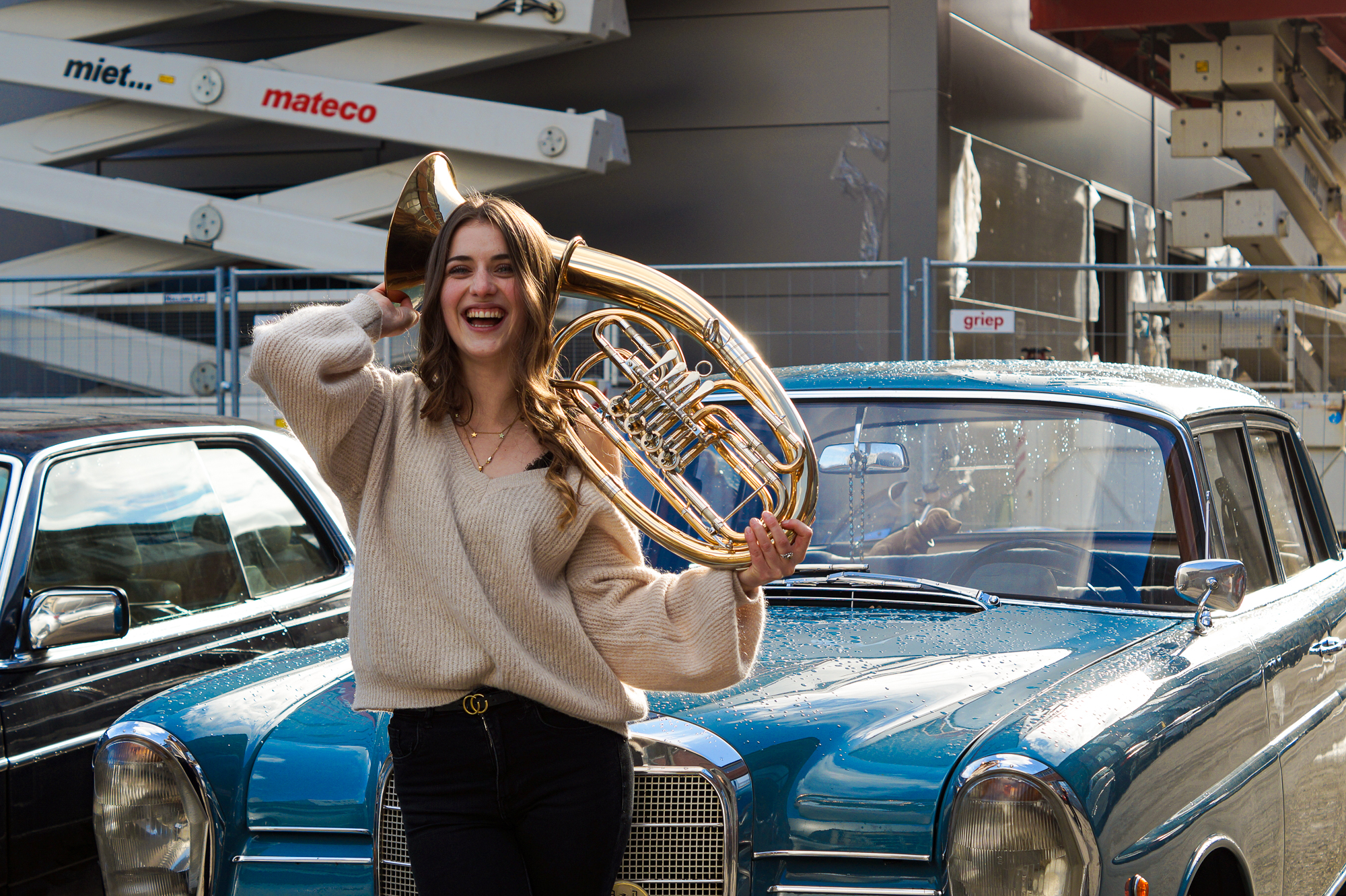 Horn player with her instrument in front of vintage car