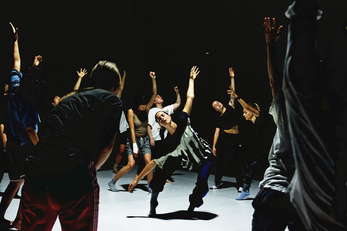 Eine Gruppe von Menschen, die mit ausladenden Bewegungen und geschlossenen Augen in einem dunklen Raum tanzen.