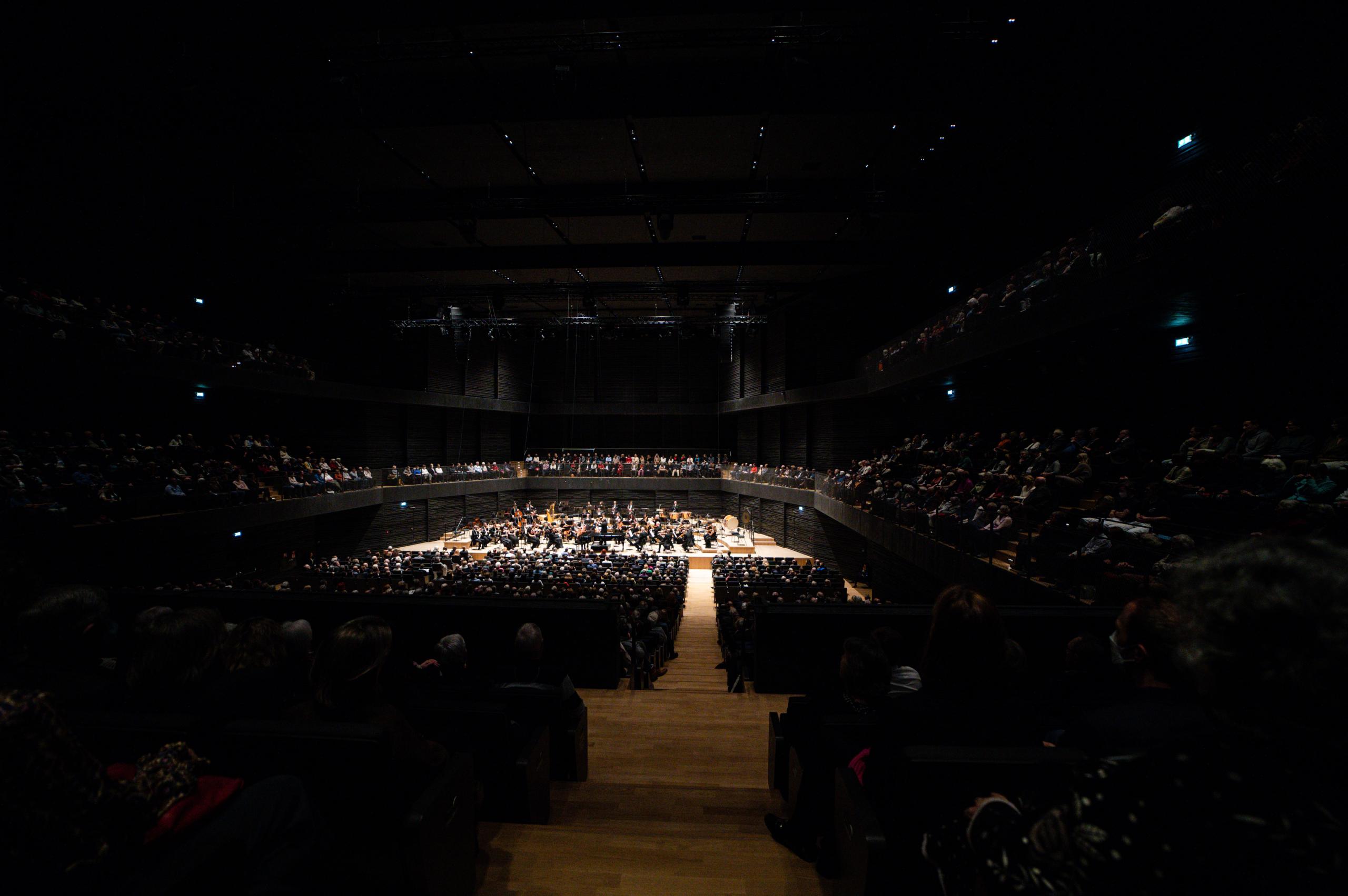 Ein dunkler Konzertsaal, der Blick geht über das Publikum hinweg von hinten auf die Bühne mit Orchester