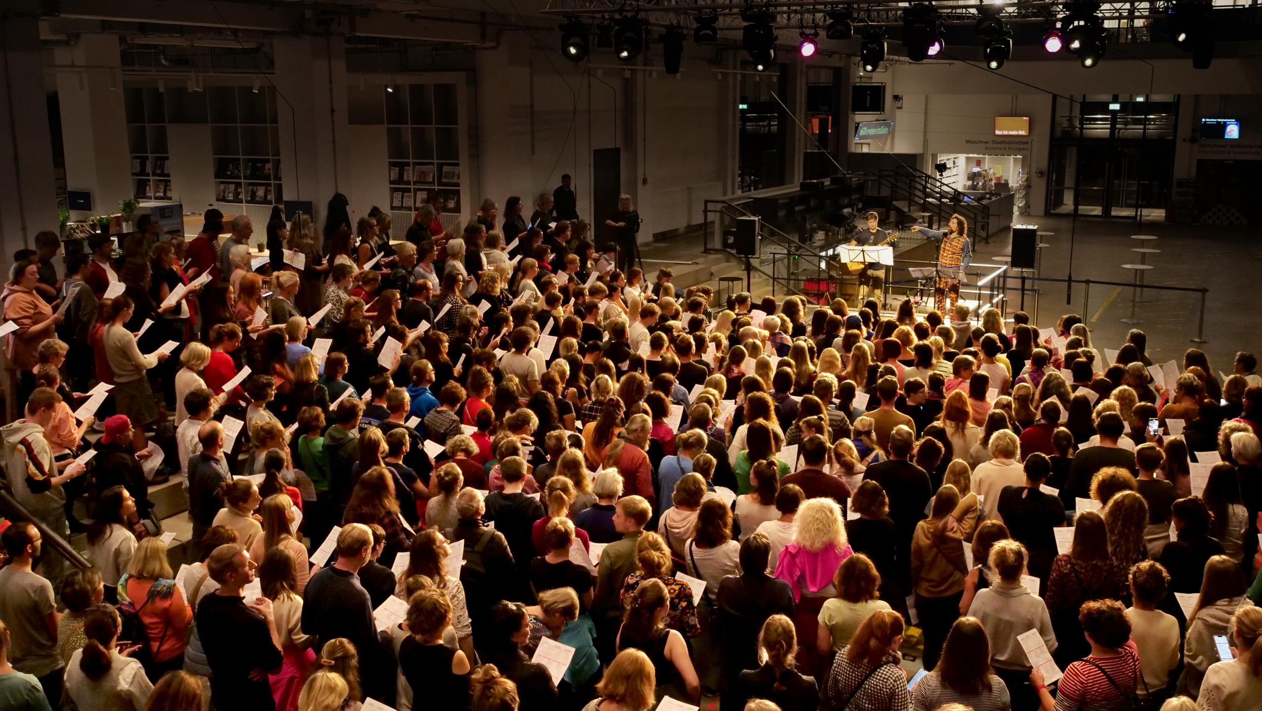 A choir sings in an industrial hall, guided by two musicians on stage.