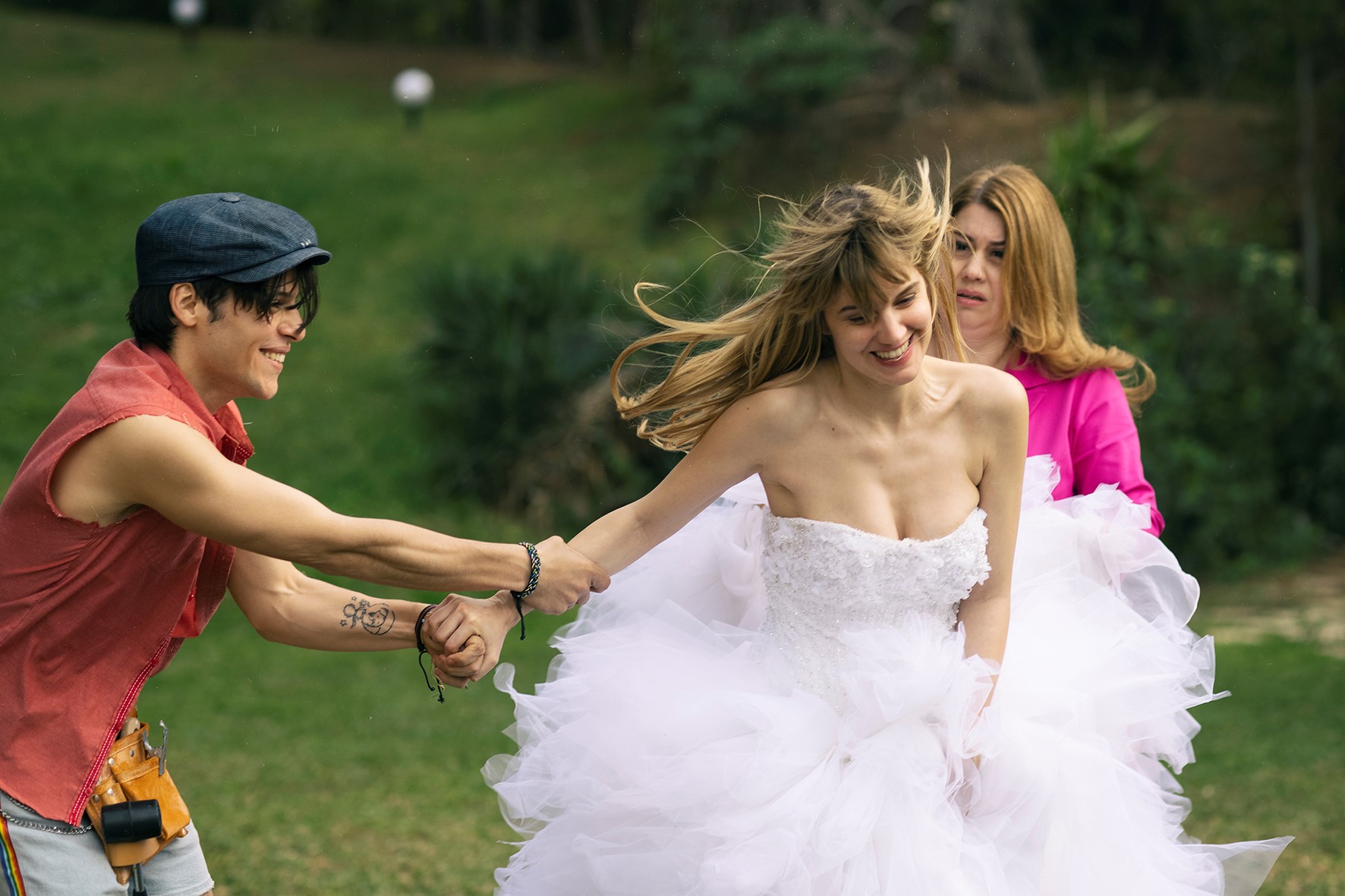 Eine junge Frau im Brautkleid scheint laufen zu wollen, wird aber von einem jungen Mann am Arm festgehalten. Beide lachen dabei. Im Hintergrund ist eine weitere Frau, die jedoch ernst guckt.