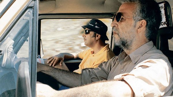 Two men wearing sunglasses are riding together in a car. The man in the passenger seat has his leg resting on the dashboard.