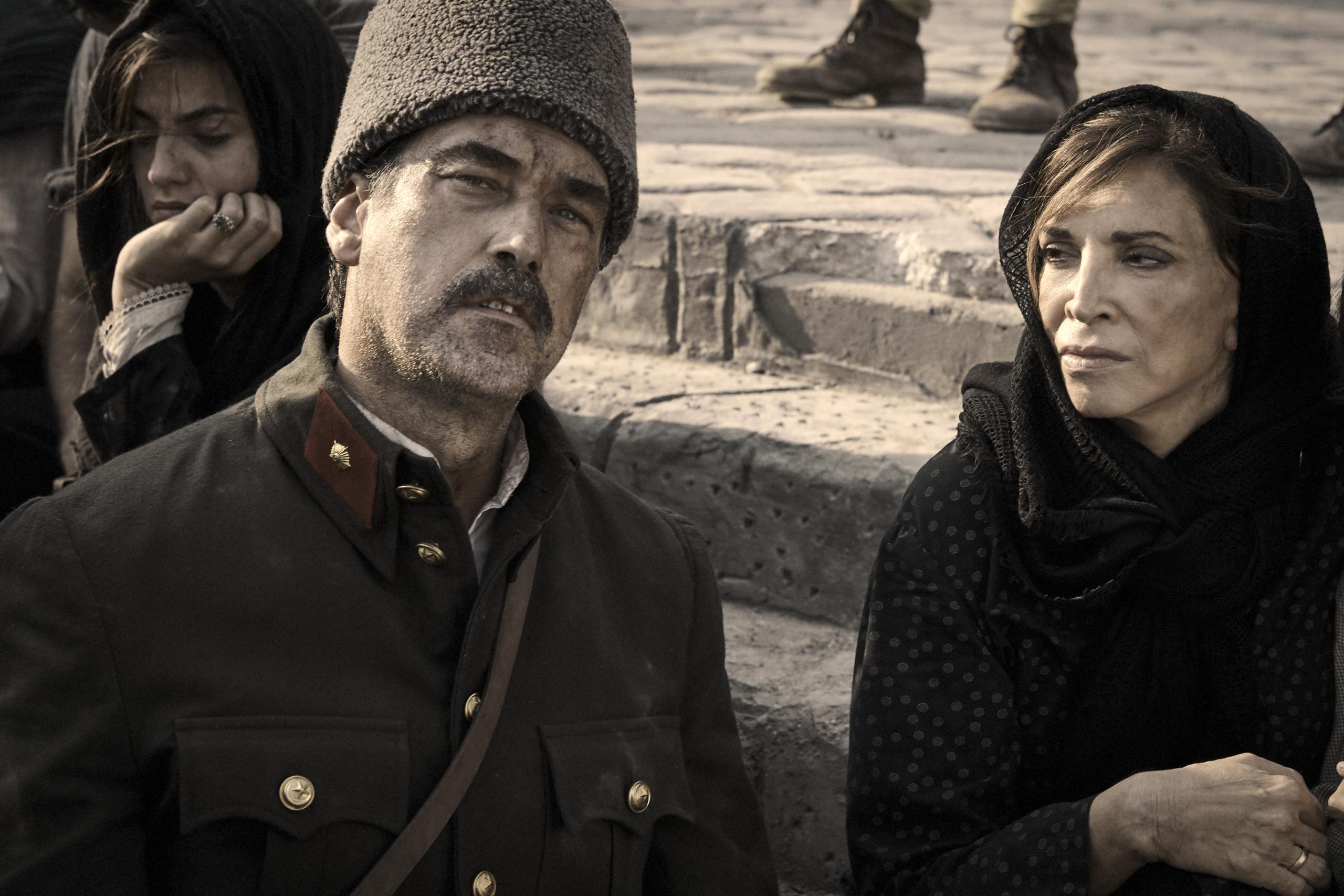 A man in military dress and a woman in a dark dress and black headscarf are sitting on stone steps with serious expressions.