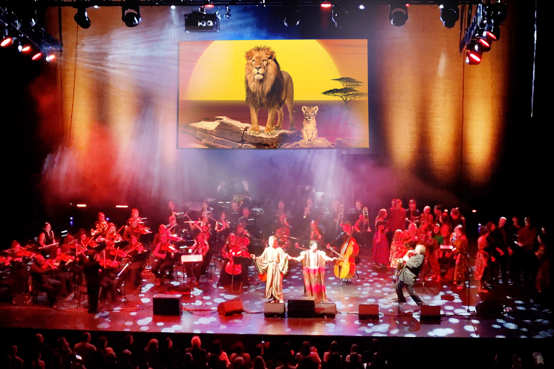 A colourfully lit stage with orchestra and singers. In the background a screen on which a lion can be seen.