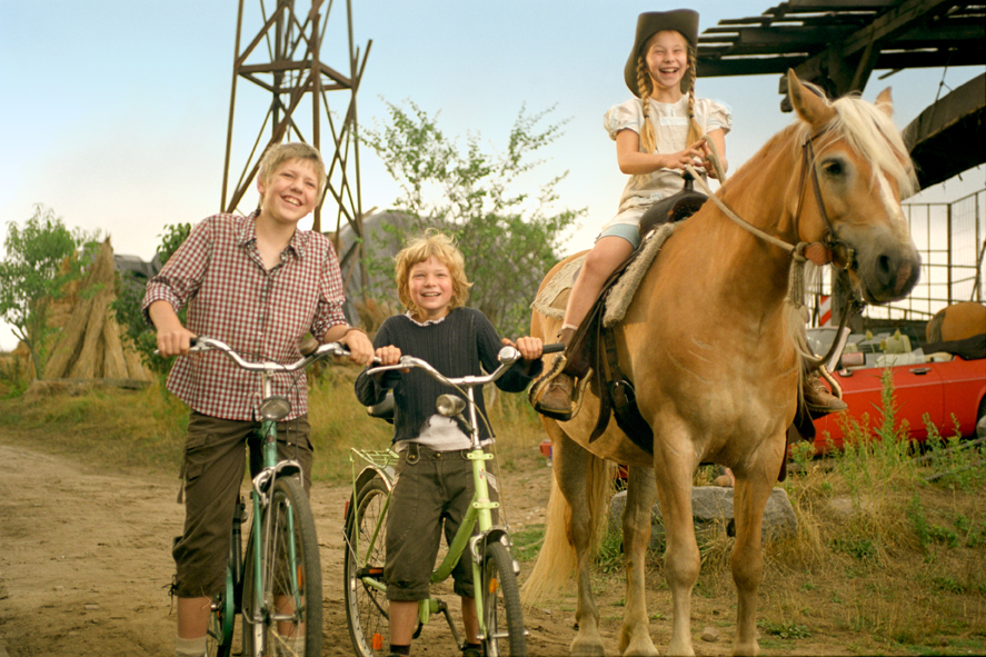 Ein Mädchen sitzt auf einem Pferd. Daneben stehen zwei Jungs mit ihren Fahrrädern.