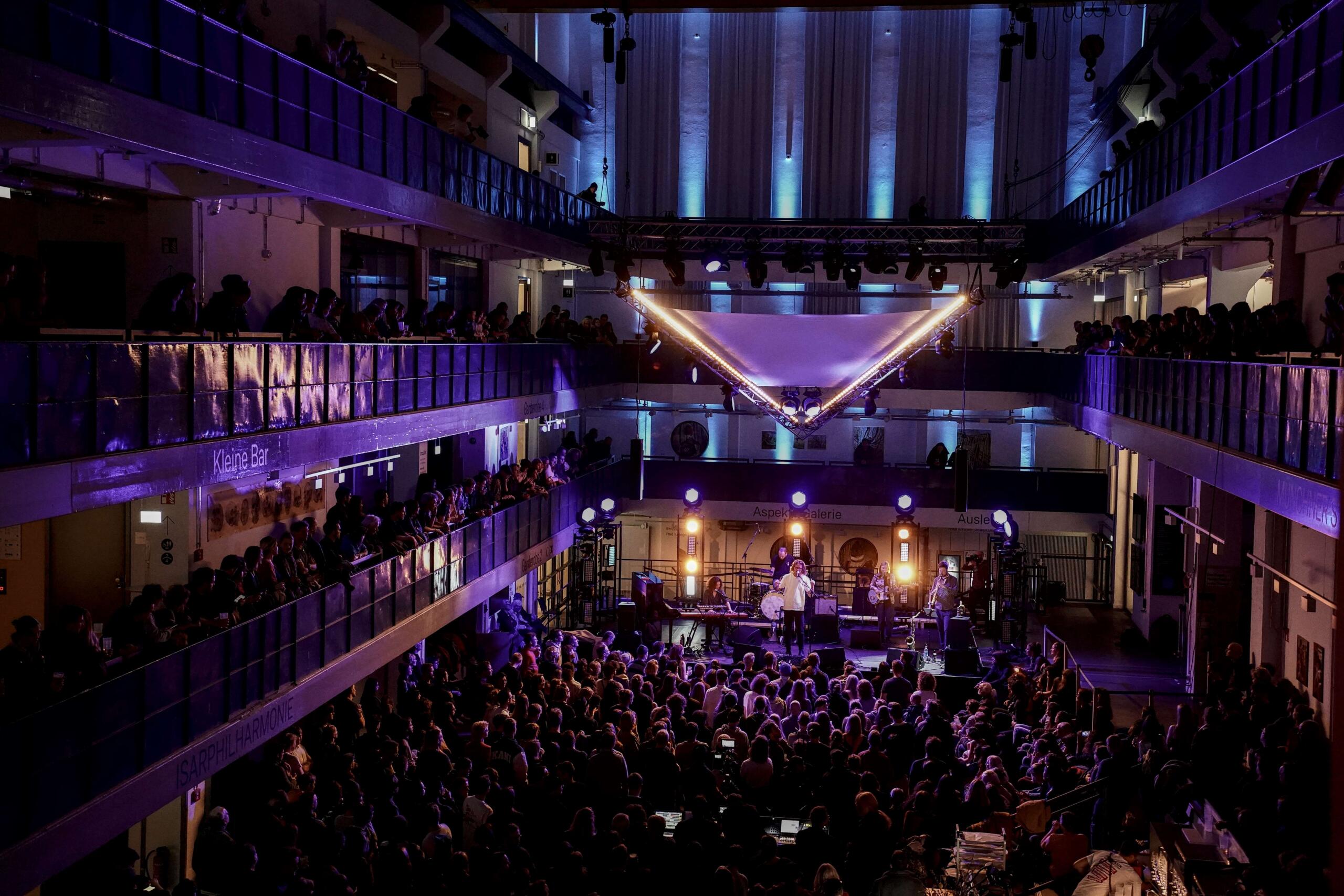 Die Halle E ist lila beleuchtet. Ein Sänger steht auf einer Bühne, Zuschauer und Zuschauerinnen  drängen sich im Erdgeschoss und auf den Balkonen.
