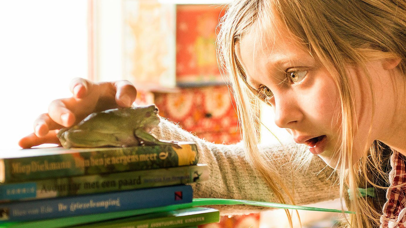 Ein Mädchen schaut staunend auf einen Frosch, der auf einem Bücherstapel sitzt.