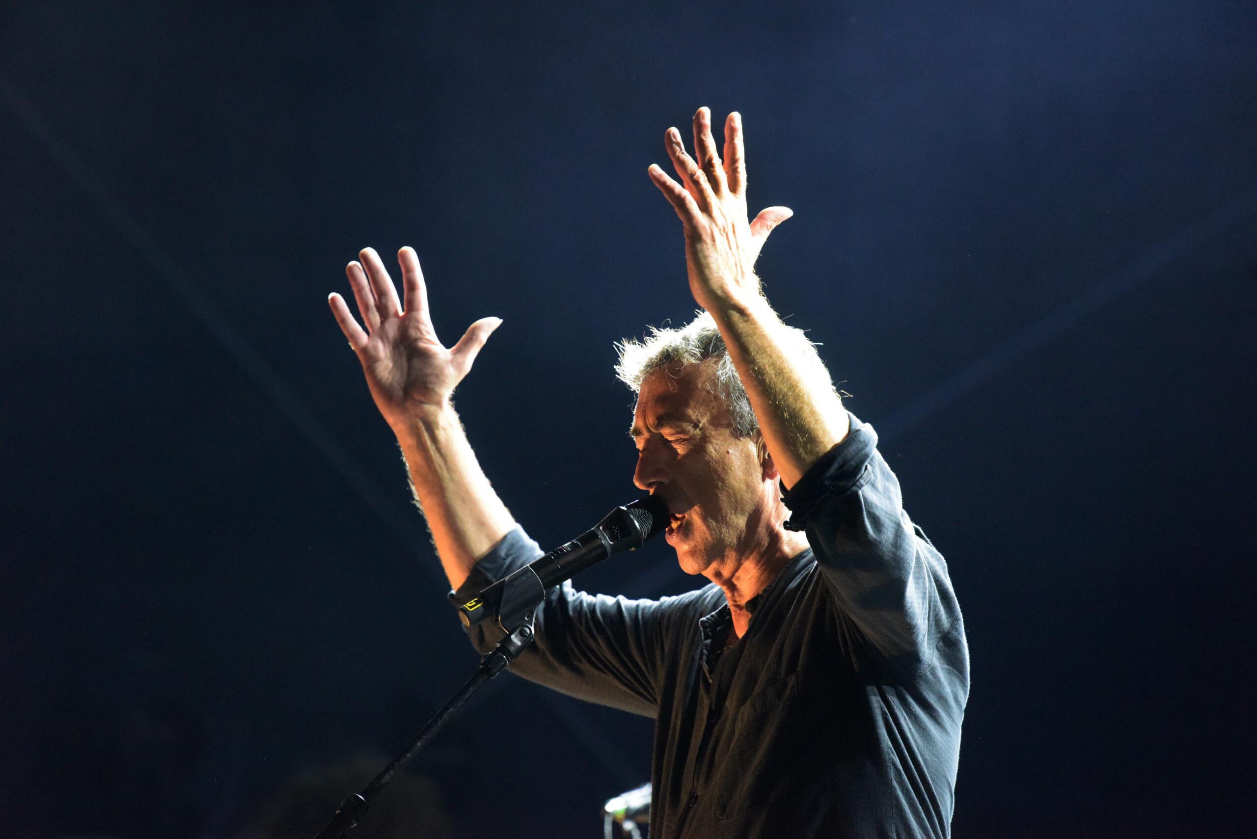 Portrait of the musician Hubert von Goisern. He stands on a stage, sings into a microphone and raises both arms.