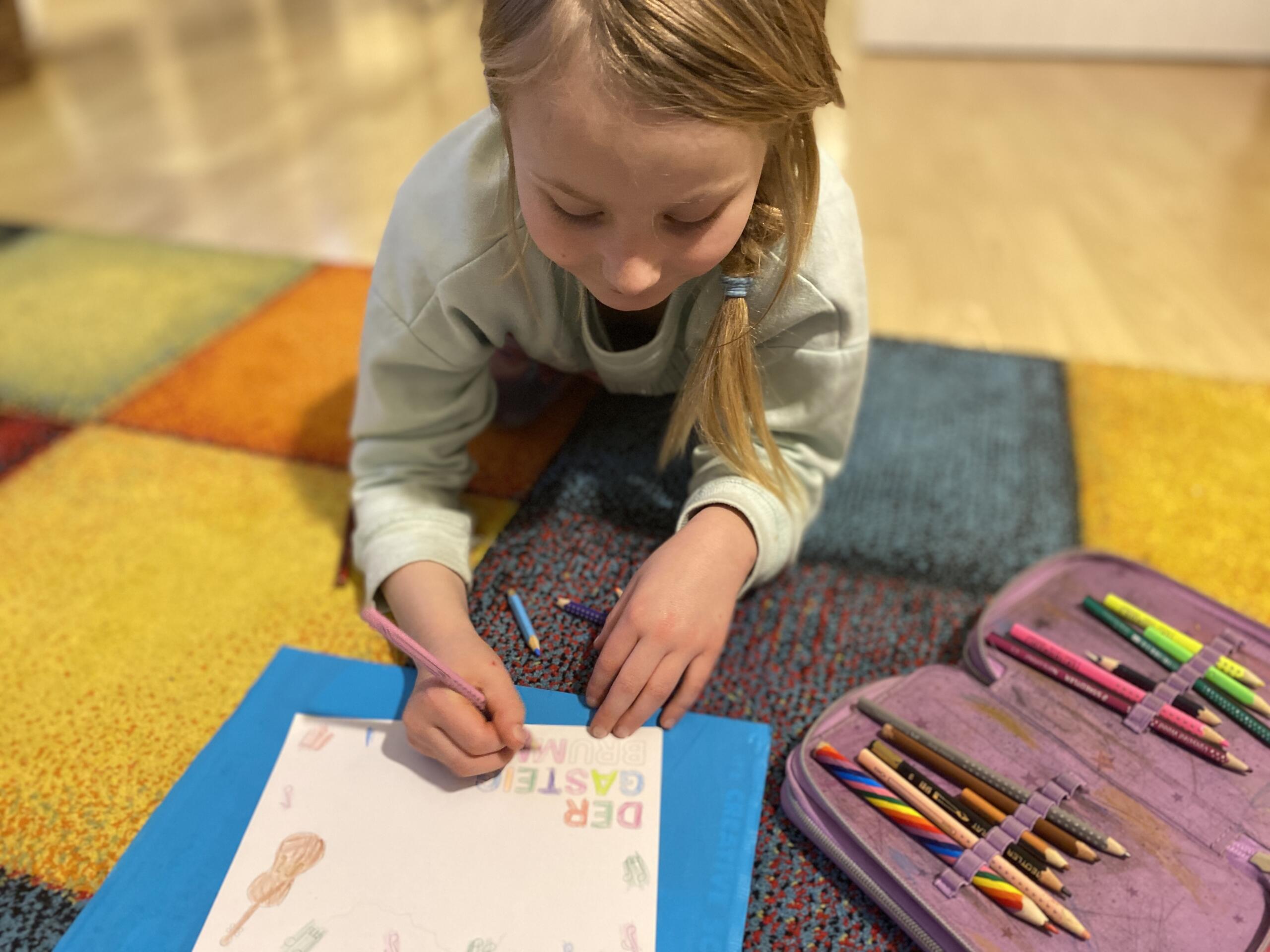 A girl lies on a colourful carpet and draws a Gasteig-brummt greetings card with coloured pencils.