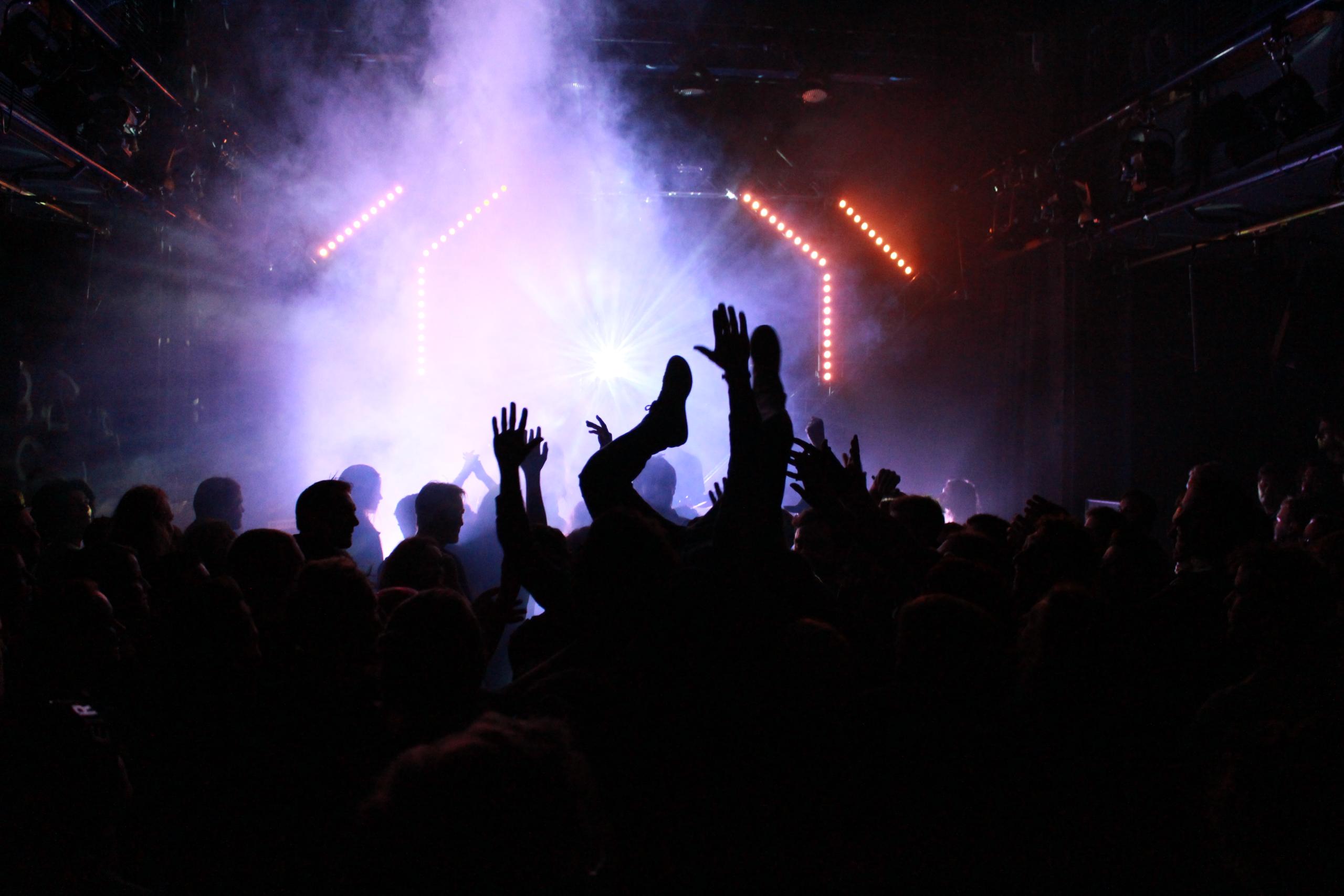 Silhouettes of audience in front of a brightly lit stage, a musician does stage diving and is carried by the audience, his legs pointing upwards.