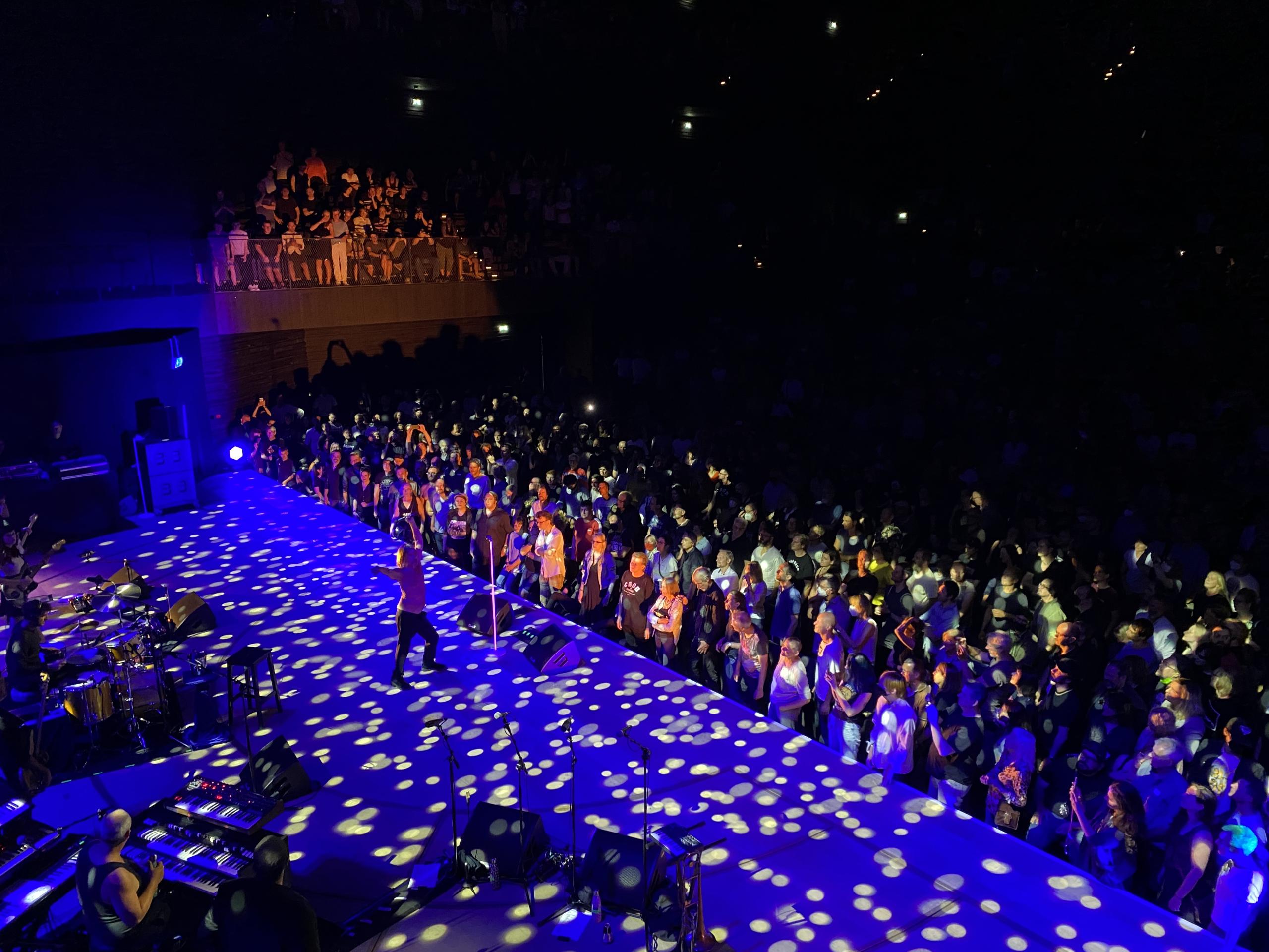 Blick von der schön beleuchteten Bühne der Isarphilharmonie. Iggy Pop steht am Rand zum Publikum