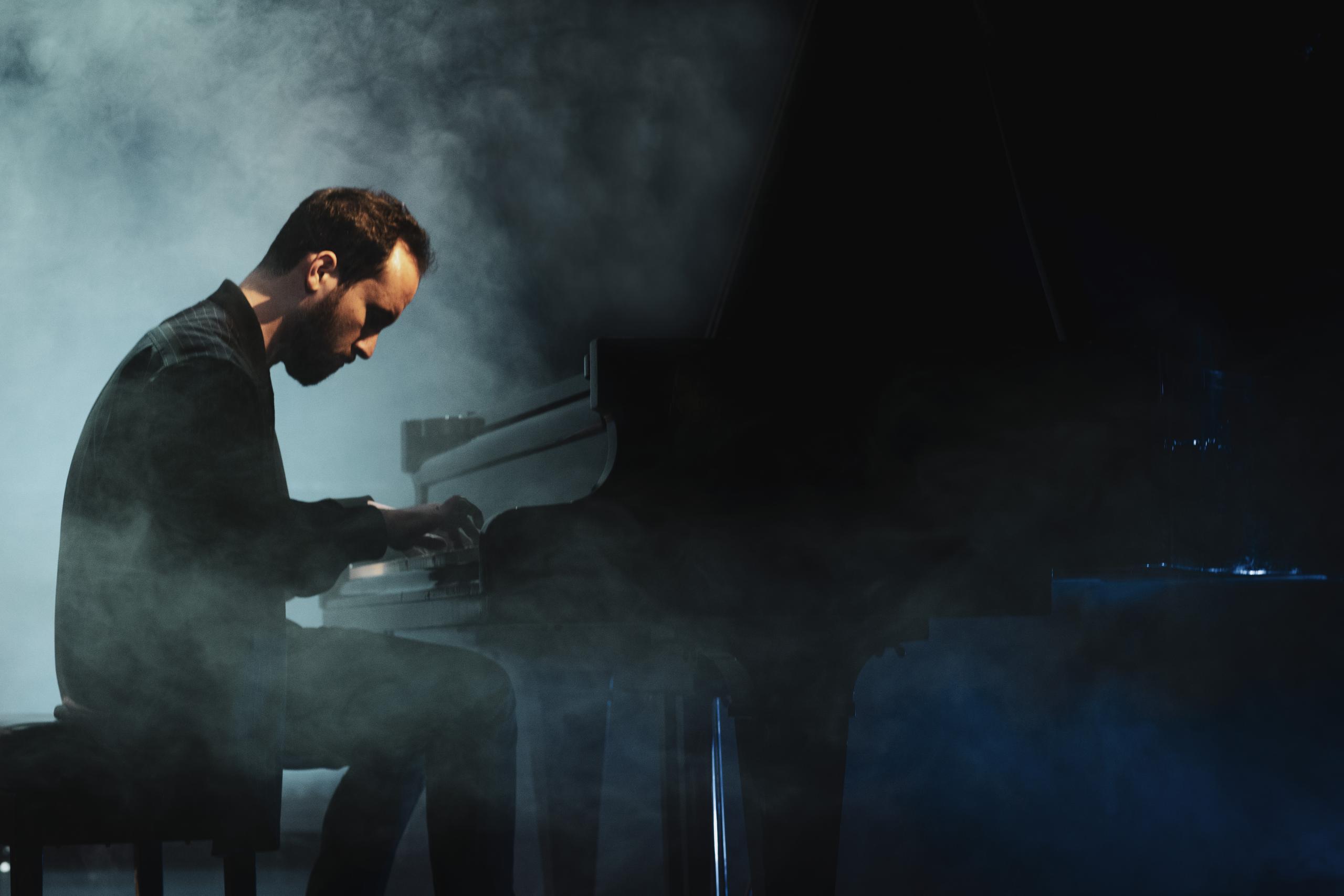 The pianist Igor Levit on the stage at the grand piano. The picture is taken from the side, around him fog.