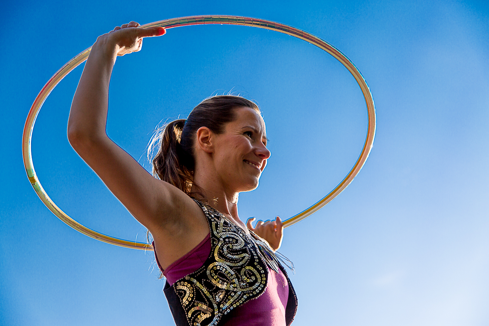 A woman against a blue sky, she holds a hula hoop over her head and beams.