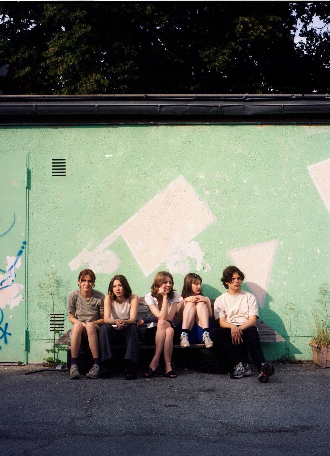 Four young women and a young man sit on a bench in front of a wall with graffiti