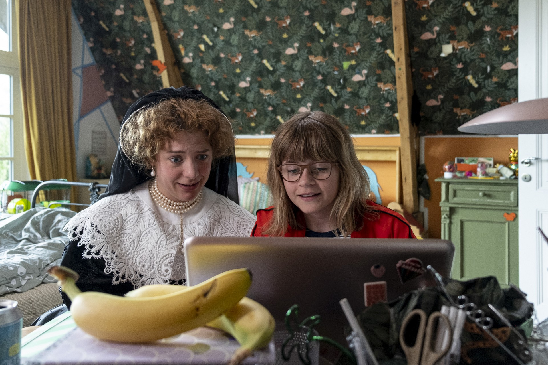 Ein Mädchen und eine Frau, die aussieht, als wäre sie einem alten Gemälde entsprungen, sitzen gemeinsam vor einem Laptop.