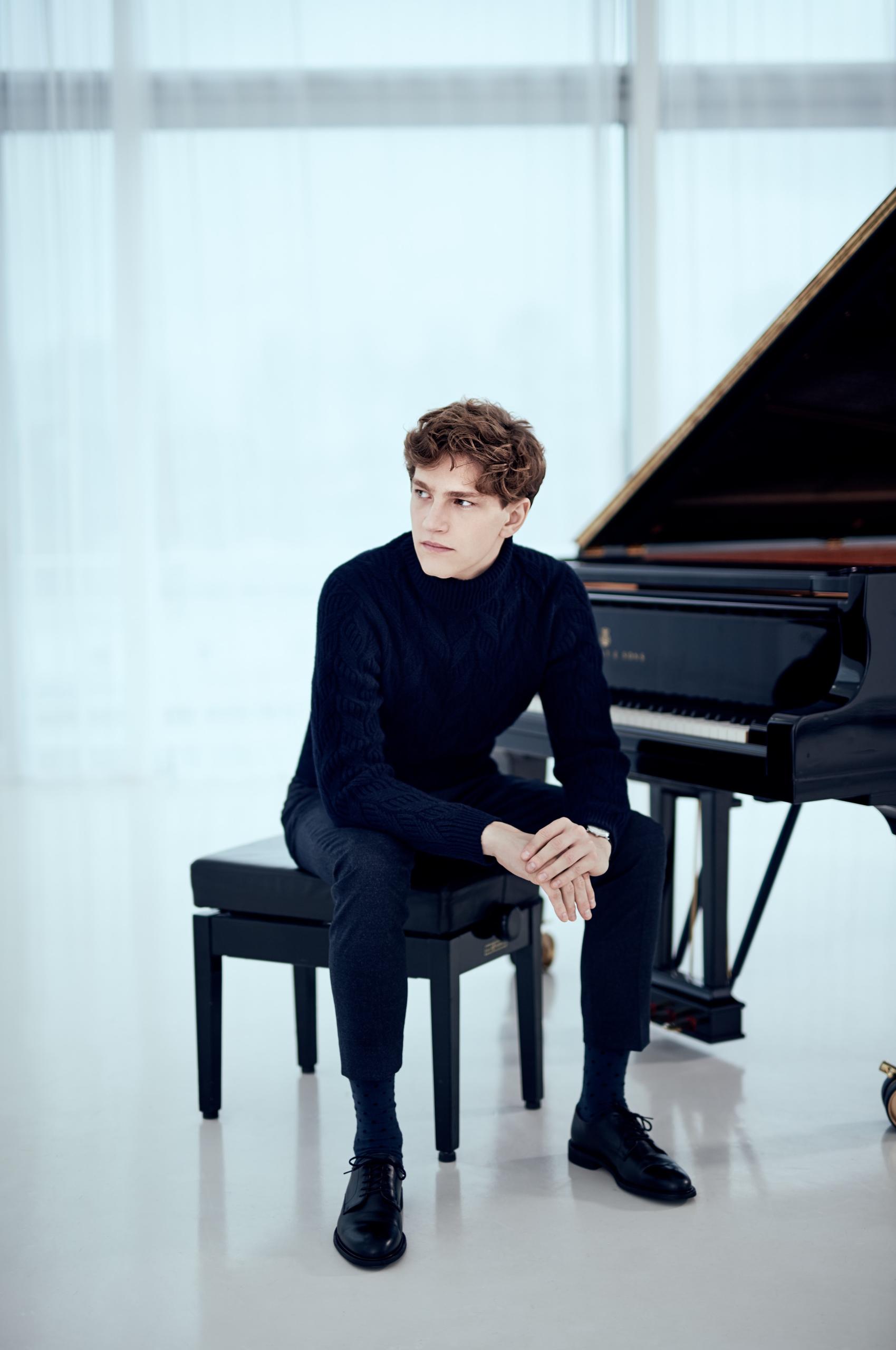 Young pianist Jan Lisiecki sits on the piano stool in front of a black grand piano