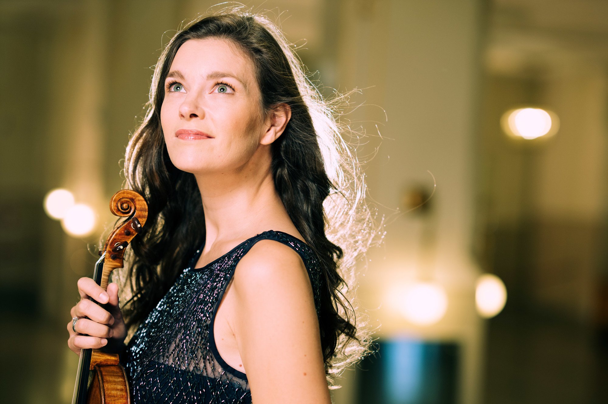 Portraitaufnahme der Geigerin Janine Jansen. Sie steht vor einem strahlendem Licht und hält ihre Geige vor sich in der Hand.