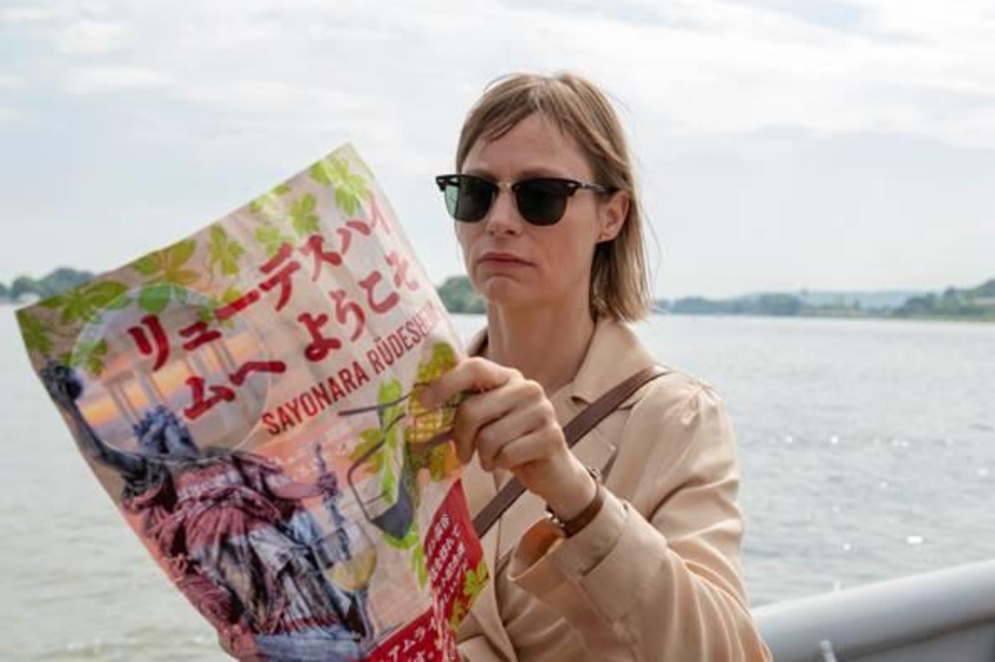 Eine Frau steht an einem großen Fluss, sie trägt eine große Sonnenbrille und halt ein bunt mit deutschen und japanischen Schriftzeichen bedrucktes Plakat in den Händen.