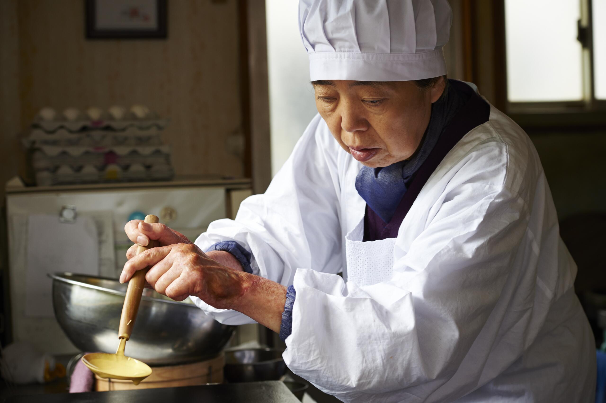 Ein japanischer Koch steht vor einem Herd und gießt Pfannkuchenteig auf eine Platte.