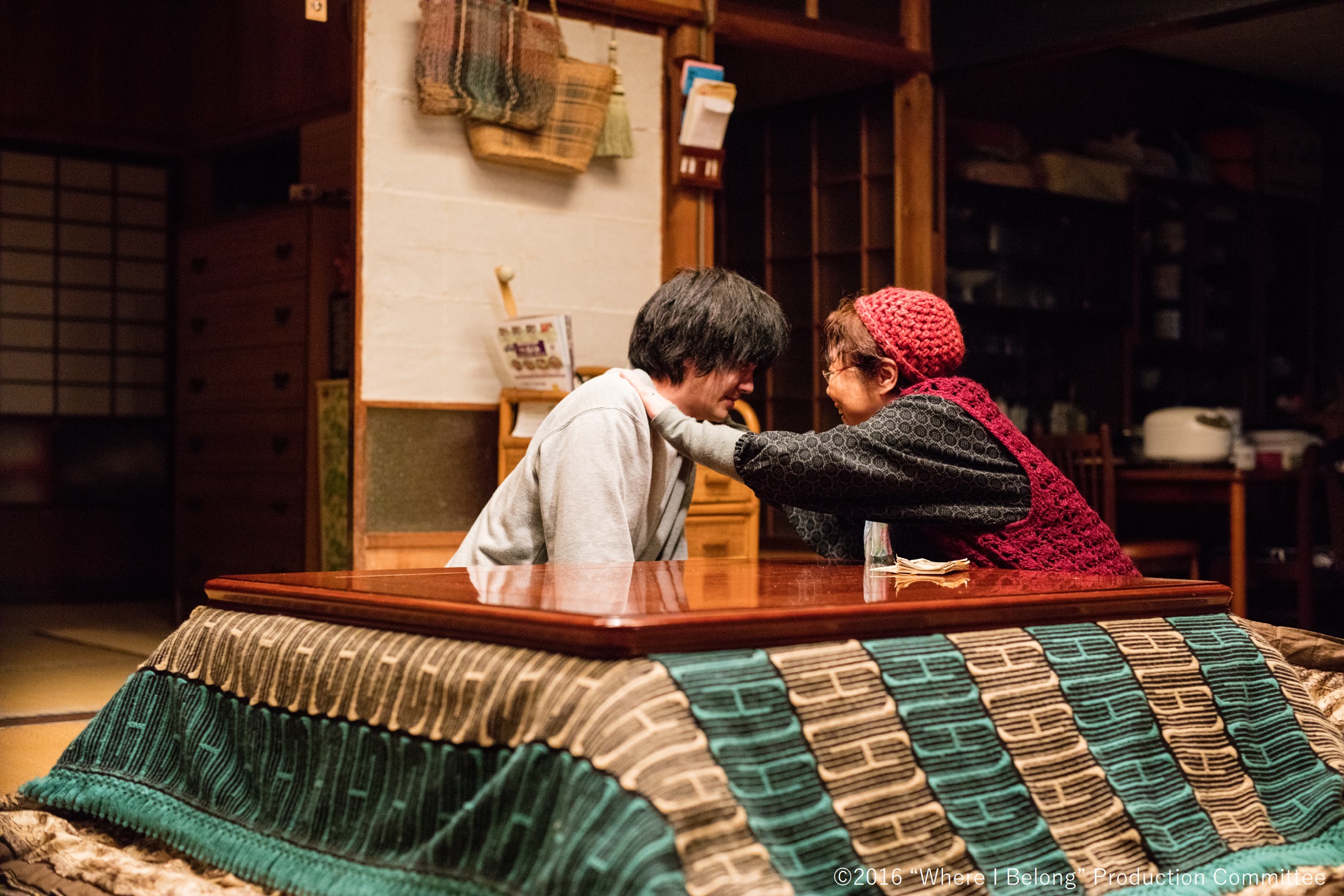 Eine alte Frau und ein jüngerer Mann sitzen hinter einem tiefgelegten Tisch auf dem Boden einer japanischen Wohnung. Sie sehen sich an und sie legt ihm die Hände auf die Schultern.