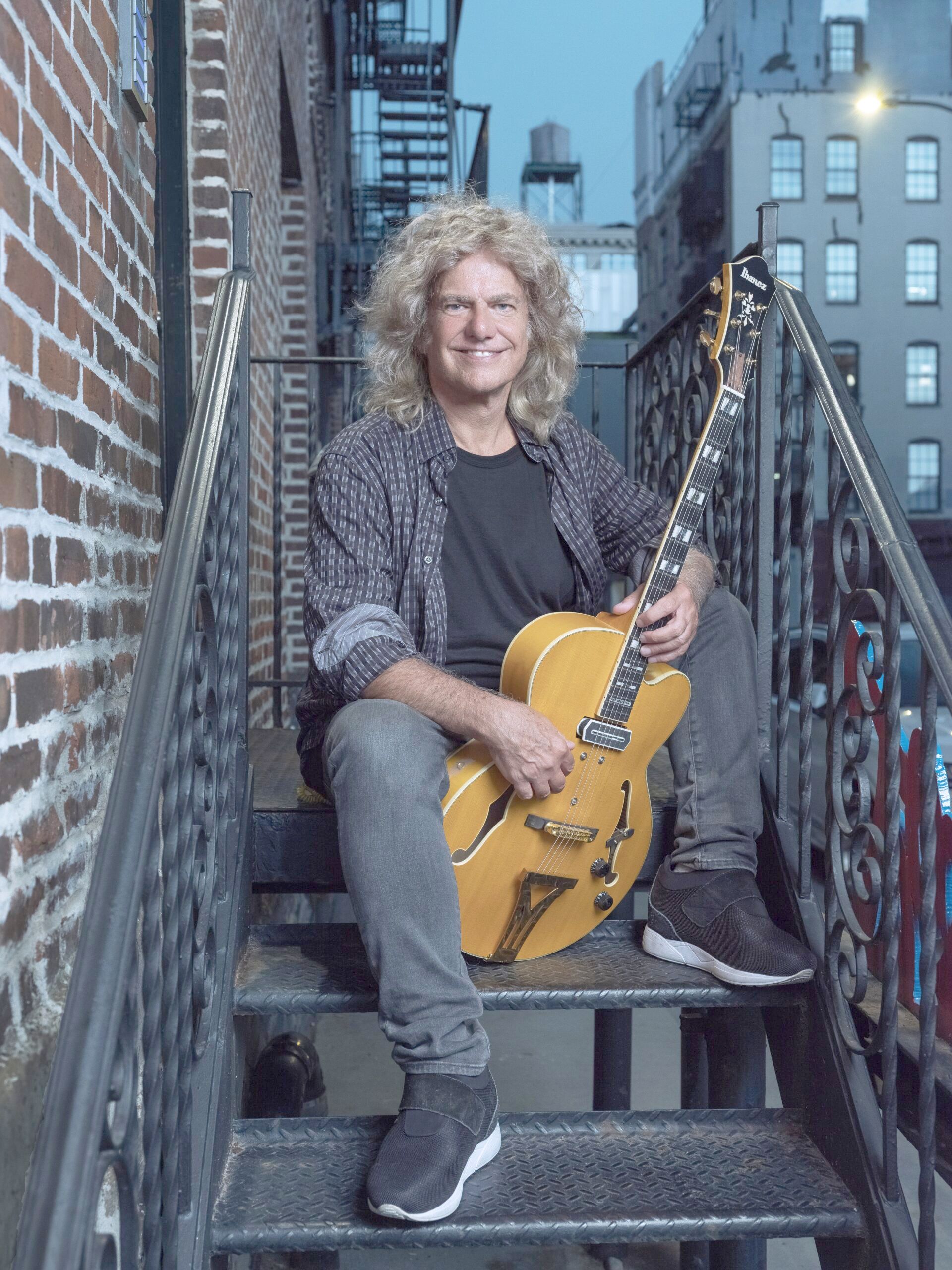 Portrait of the guitarist Pat Metheny. He sits with a guitar on a fire escape of a building and smiles into the camera.