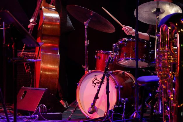 Ausschnitt eines Bühnenauftritts. Man sieht ein Schlagzeug und eine Hand des Drummers, außerdem einen Kontrabass links und rechts ein Saxophon.