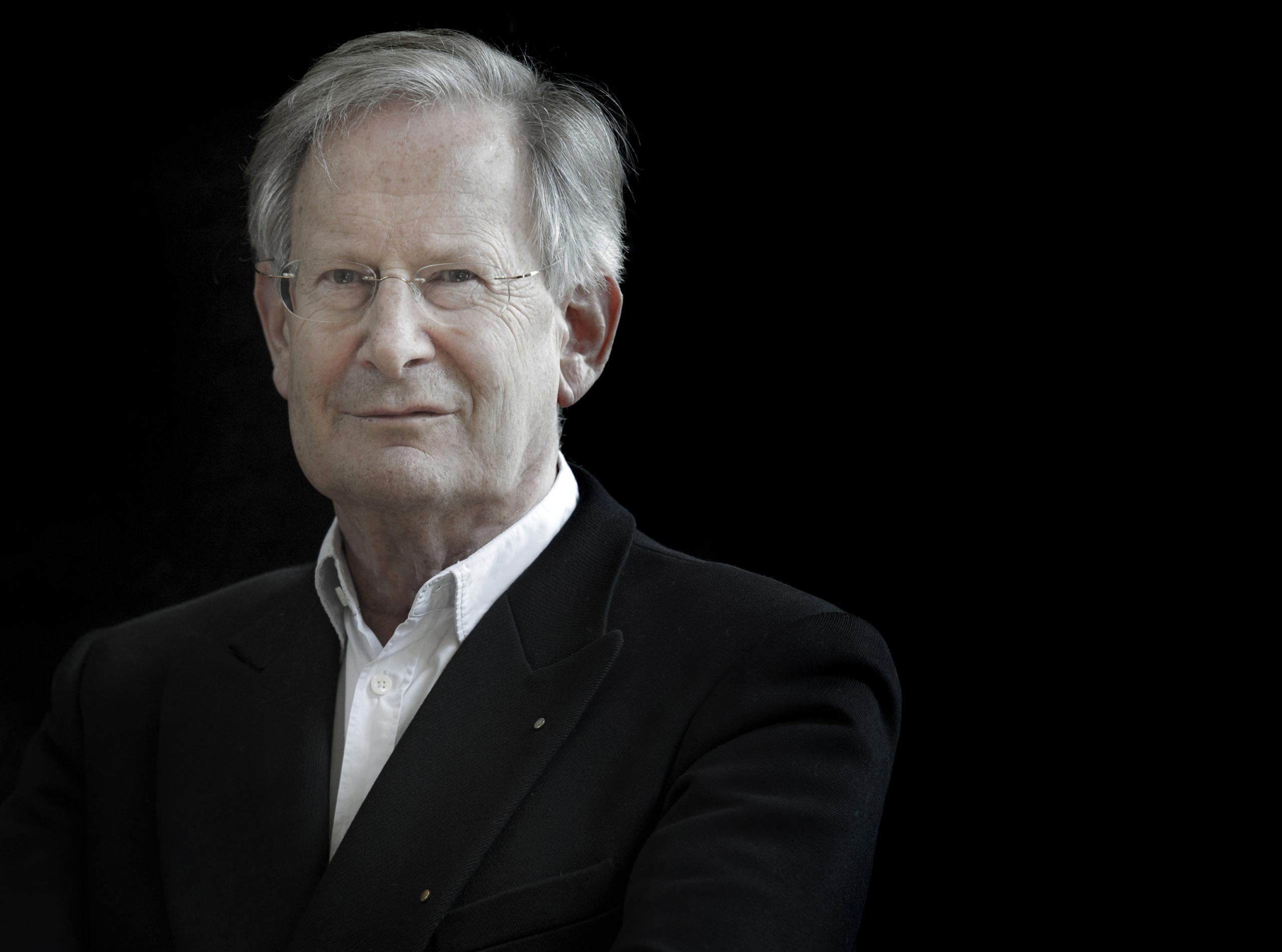 Portrait shot of conductor Sir John Eliot Gardiner smiling at the camera against a black background.